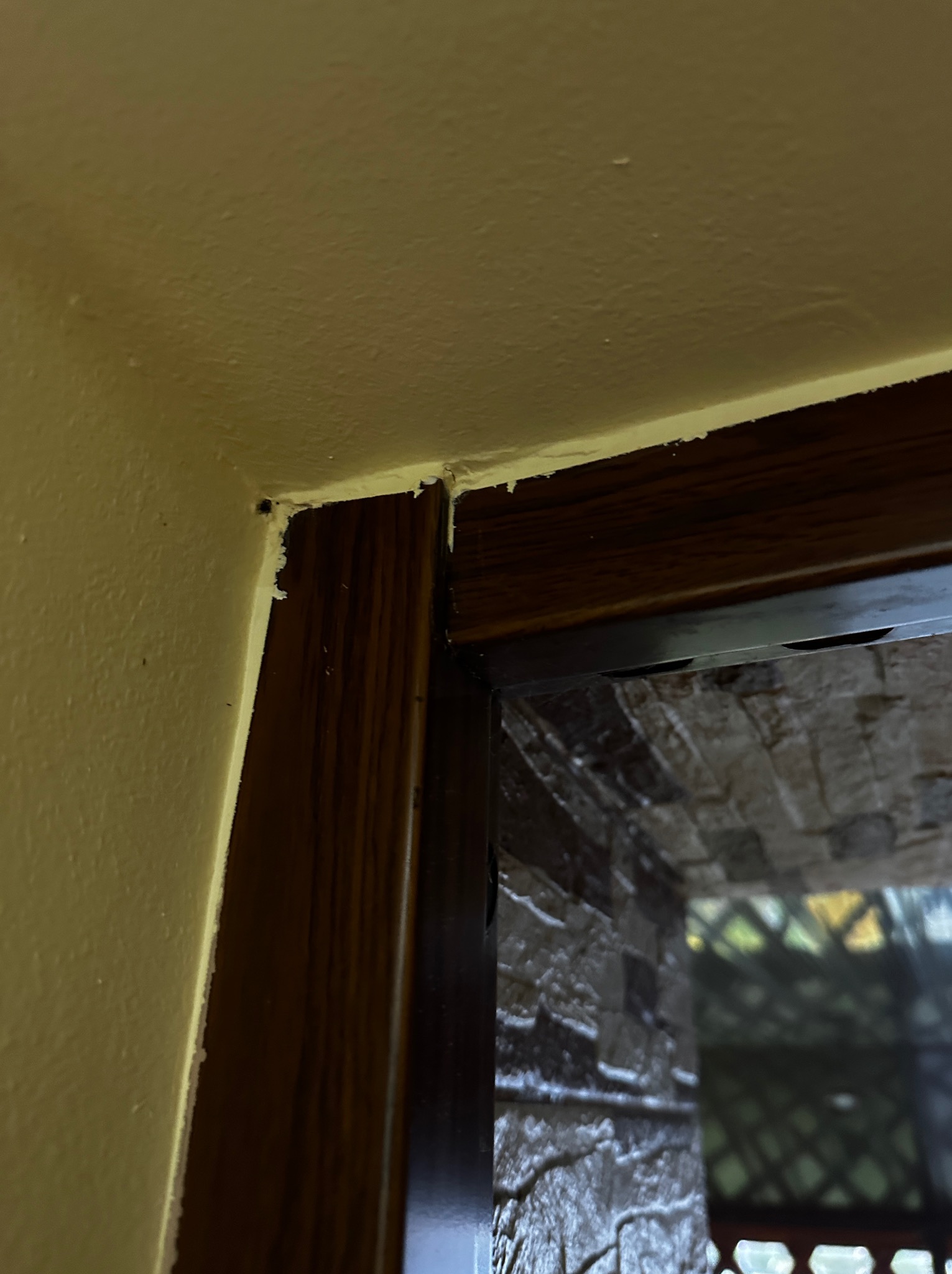 Narożnik ściany i futryny drzwiowej z widocznymi niedoskonałościami wykończenia, ukazujący detale łączenia elementów i fakturę ściany. Widok na zewnątrz przez drzwi.