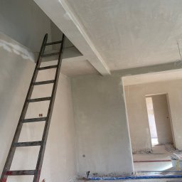 Jakub kaczmarek - Świeżo położone, jasne tynki na ścianach i suficie w surowym wnętrzu. Widoczna drewniana drabina oparta o ścianę, kable na podłodze i wiaderka po tynku.