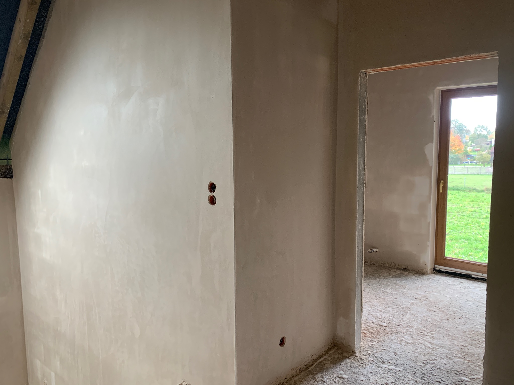 Świeżo położone, jasne tynki maszynowe na ścianach wewnątrz budynku, widoczne gniazdka elektryczne i surowy stan podłogi, okno z widokiem na zieleń.