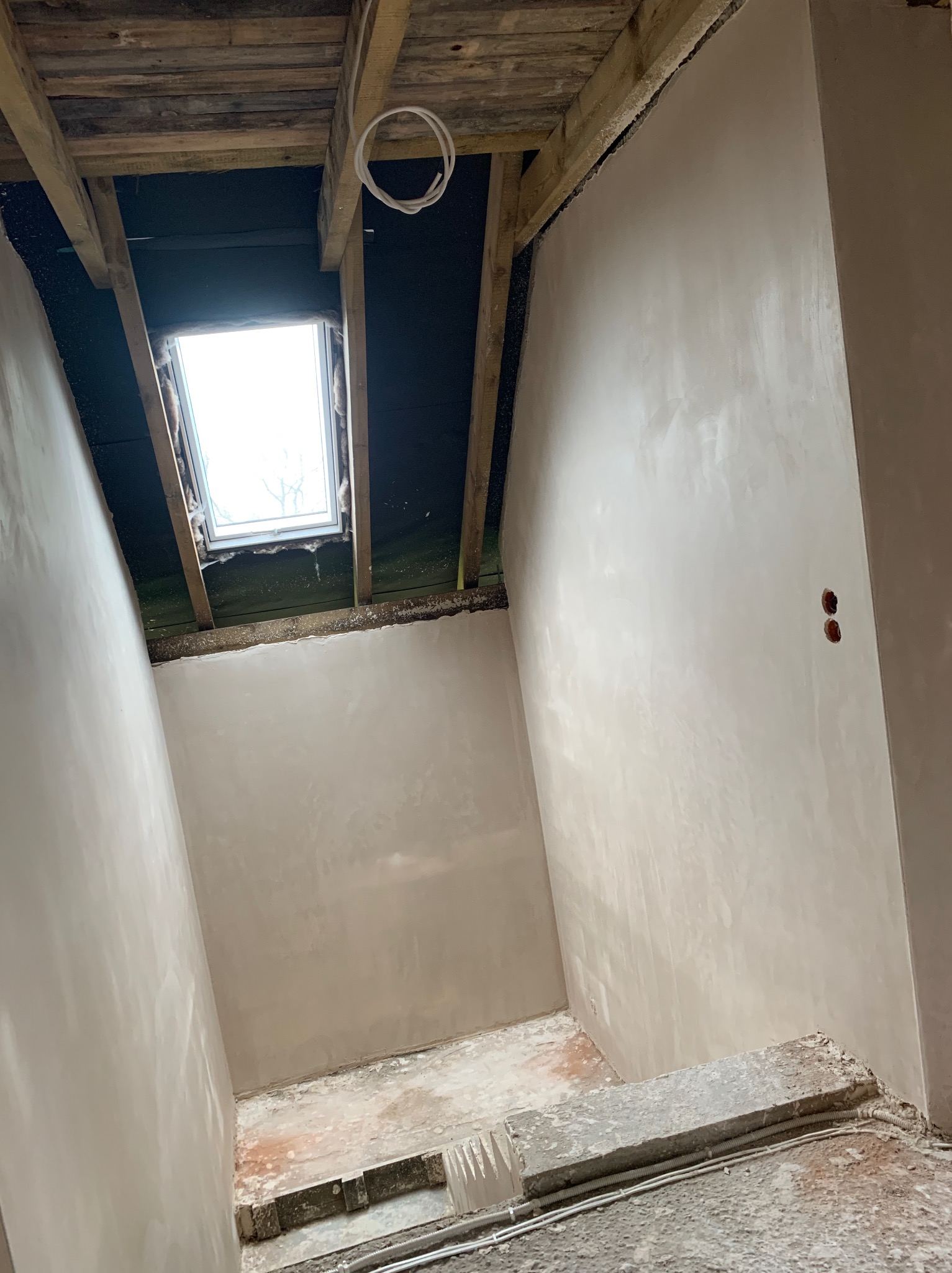 Wnętrze po tynkowaniu maszynowym: gładkie, jasne ściany, drewniany strop z oknem dachowym. Widoczne kable elektryczne i fragment podłogi w trakcie prac wykończeniowych.