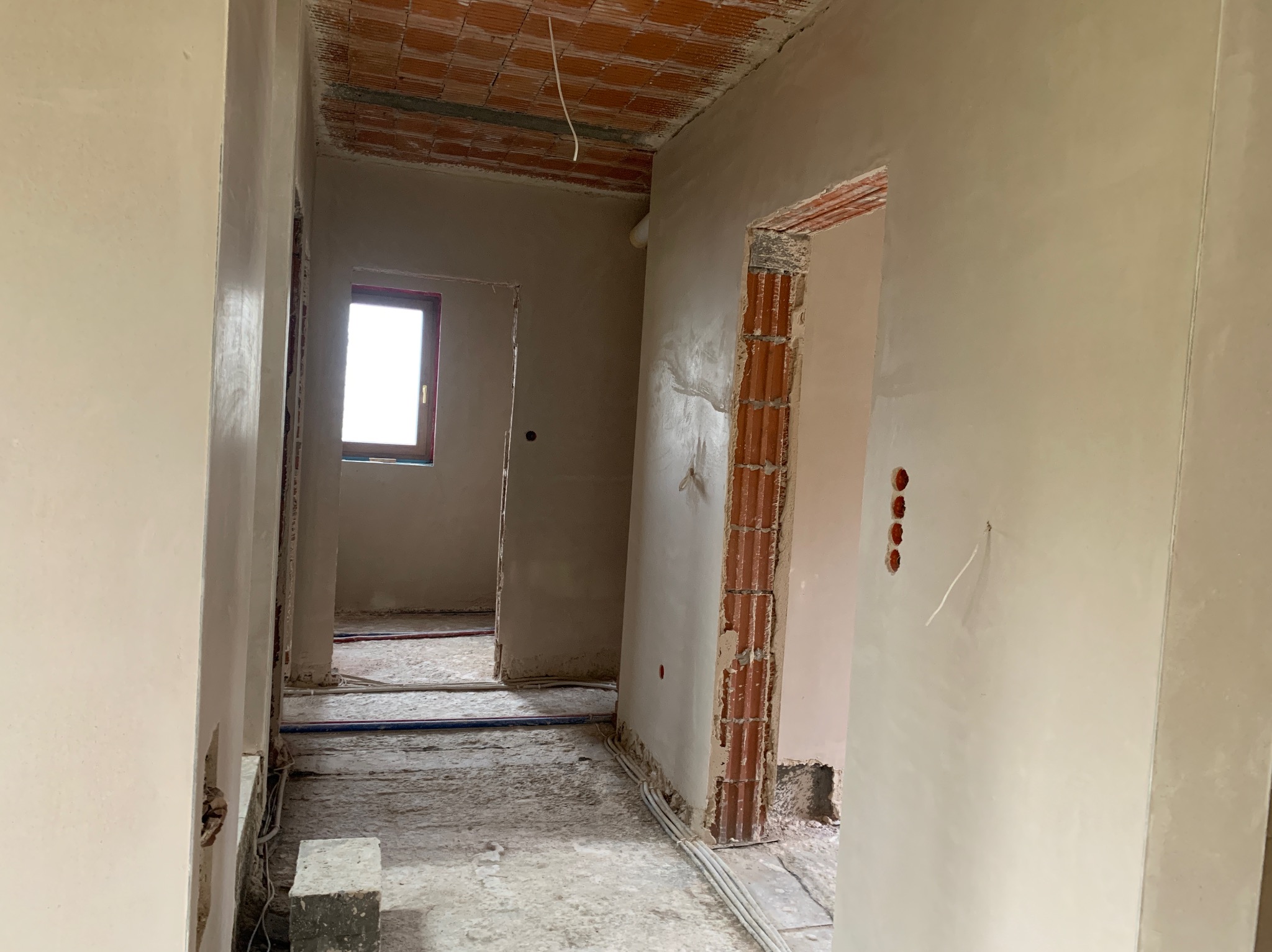 Wnętrze w trakcie prac tynkarskich: świeżo otynkowane ściany w jasnym odcieniu, widoczne cegły wokół otworów drzwiowych, kable na podłodze i surowy strop.