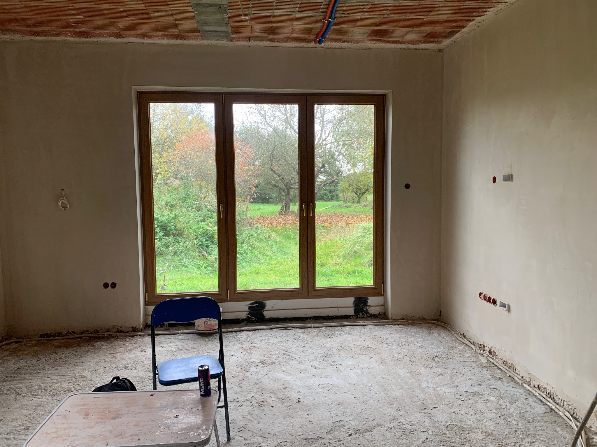 Wnętrze pokoju po nałożeniu tynków, widoczne gładkie ściany, okno z widokiem na ogród, instalacje elektryczne i fragmenty wyposażenia budowlanego.