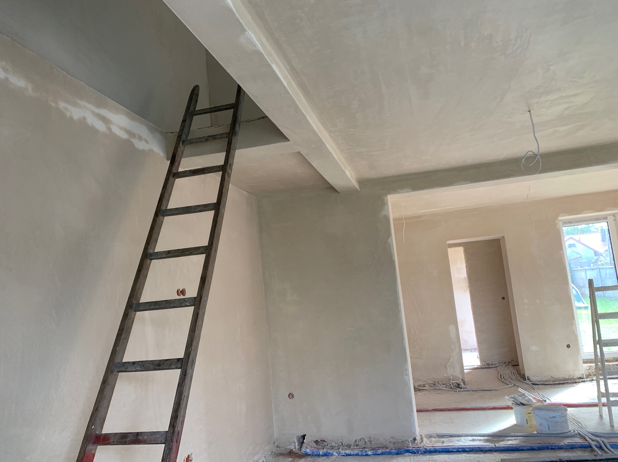 Świeżo położone, jasne tynki na ścianach i suficie w surowym wnętrzu. Widoczna drewniana drabina oparta o ścianę, kable na podłodze i wiaderka po tynku.