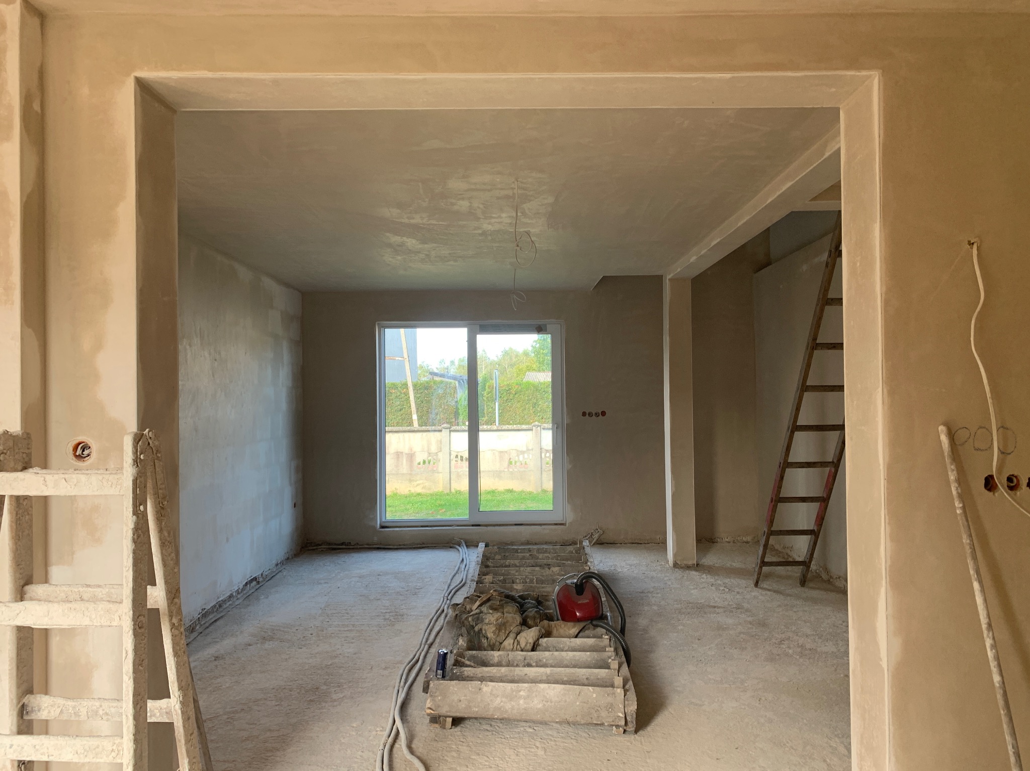 Wnętrze domu w trakcie prac tynkarskich: ściany pokryte świeżym tynkiem, widoczne drabiny, przewody elektryczne i okno z widokiem na zieleń. Jasne, naturalne światło.