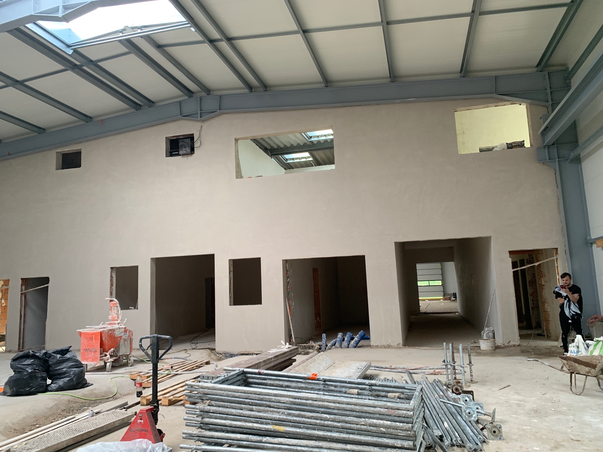 Wnętrze hali po nałożeniu tynków: gładkie ściany, widoczne otwory okienne i drzwiowe, sprzęt budowlany, stalowa konstrukcja dachu z oknami dachowymi.