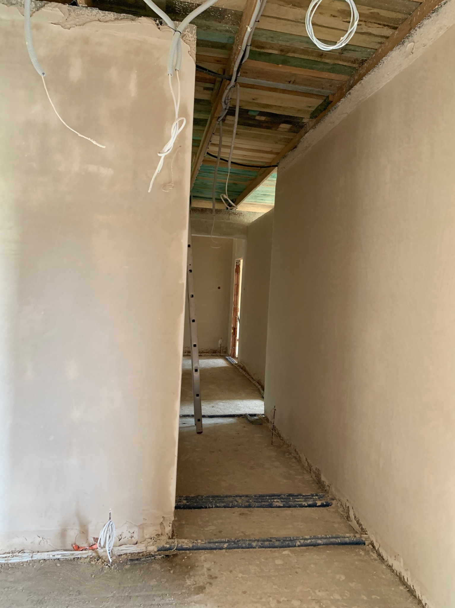 Wnętrze w trakcie tynkowania maszynowego: świeżo otynkowane ściany, widoczne przewody elektryczne i drabina w przejściu. Surowy stan wykończenia, przygotowanie do dalszych prac.