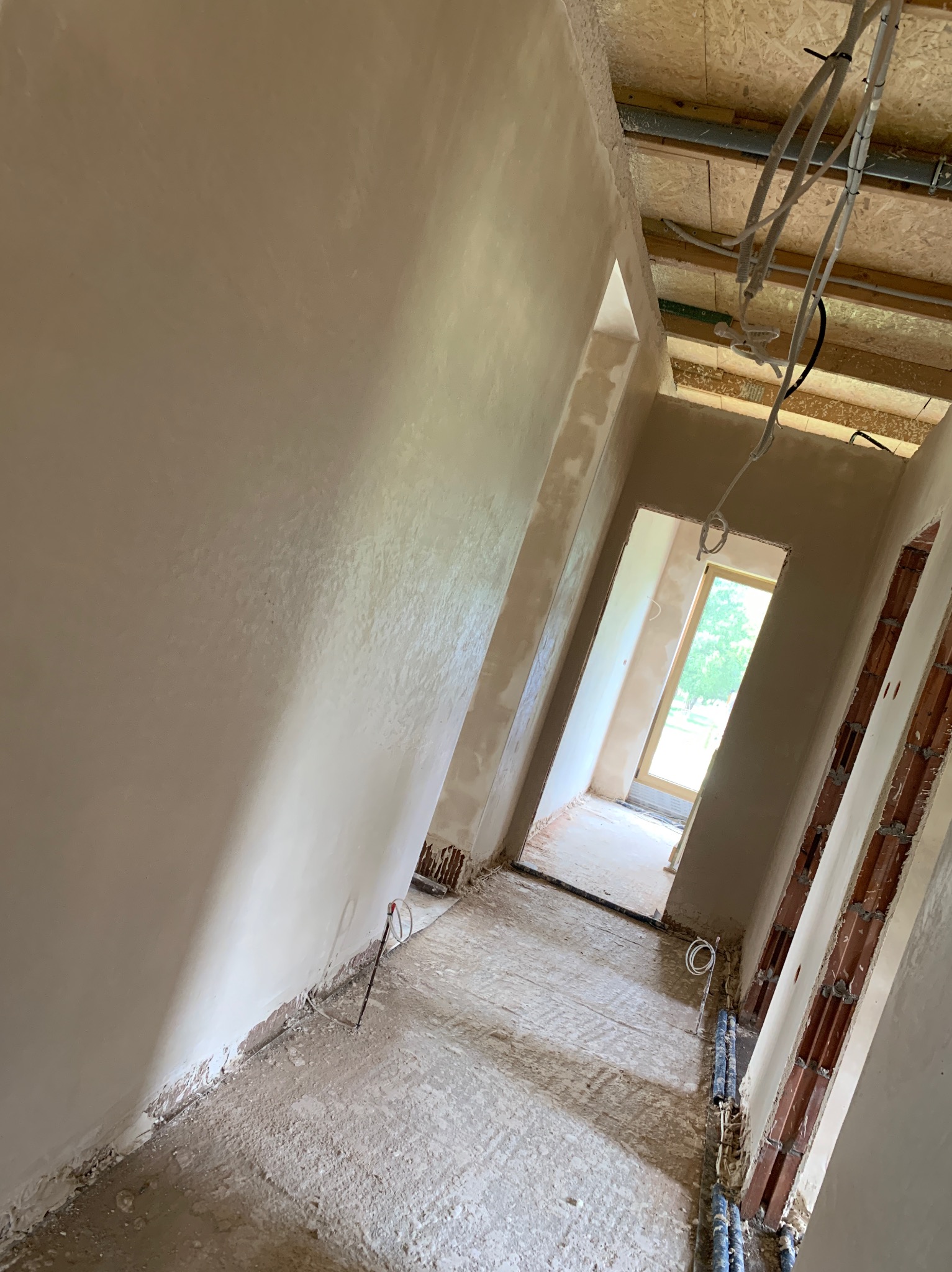 Wnętrze w trakcie prac wykończeniowych: świeżo położone, gładkie tynki na ścianach, widoczne instalacje pod sufitem i w podłodze, surowy stan podłoża.