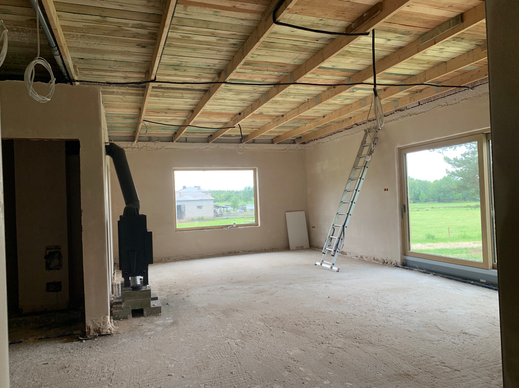 Wnętrze w trakcie prac wykończeniowych: ściany po tynkowaniu, drewniany strop, okno z widokiem na zieleń, drabina oparta o ścianę, piec wolnostojący. Surowy stan.