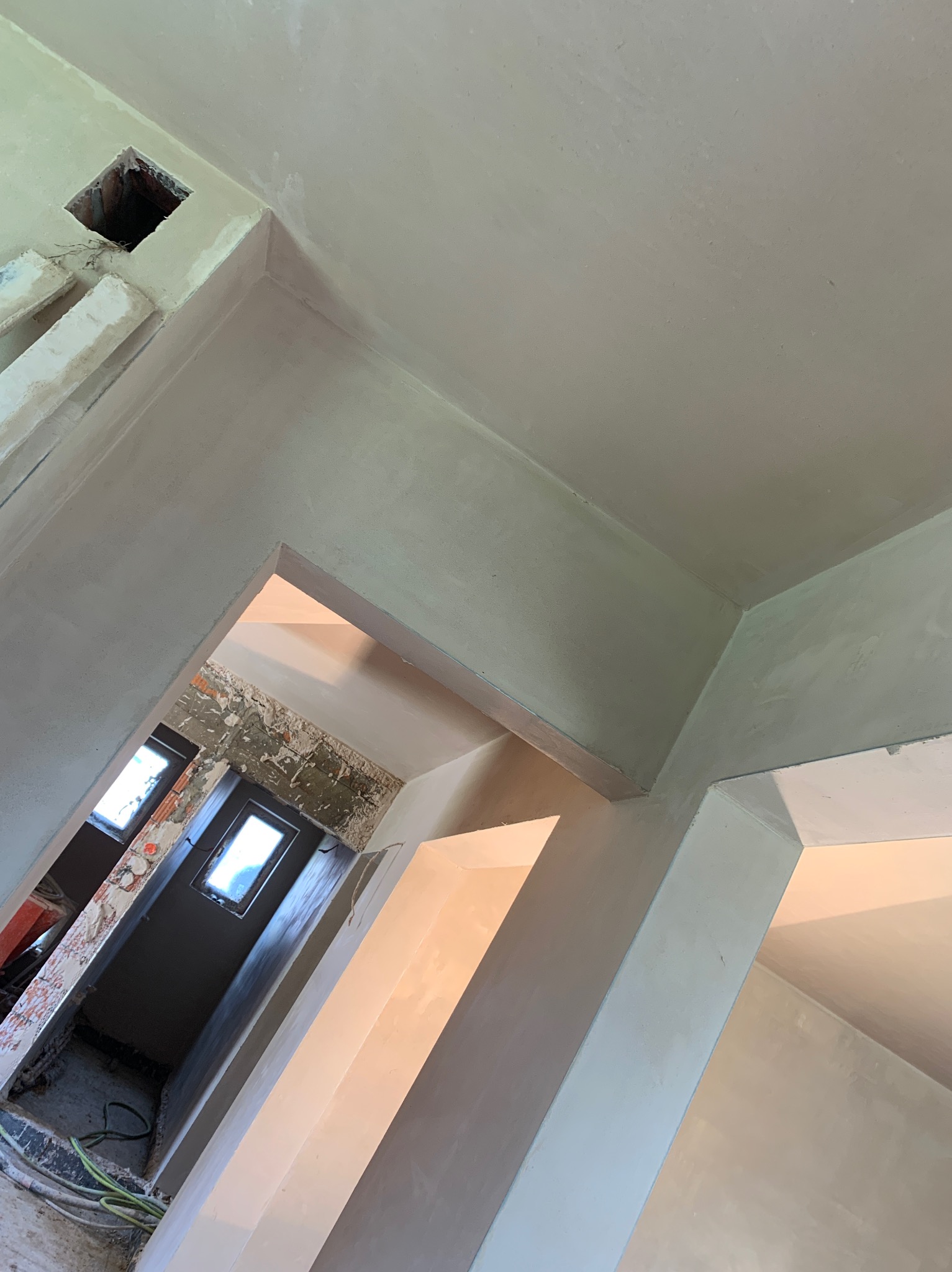 Świeżo otynkowane wnętrze z widocznymi otworami wentylacyjnymi i fragmentami surowych ścian w tle, ukazane z perspektywy sufitu.