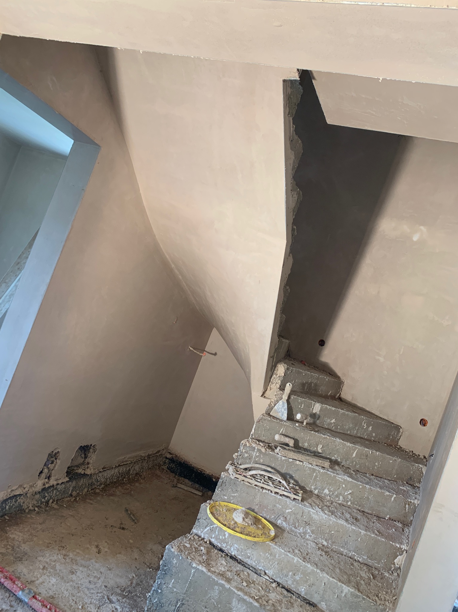 Wnętrze w trakcie prac: otynkowane ściany i sufit klatki schodowej, widoczne narzędzia murarskie na betonowych schodach. Surowy stan wykończenia.