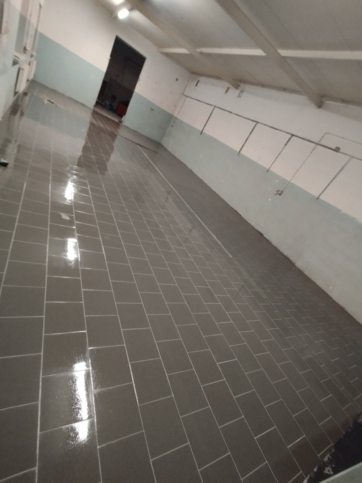 Połyskująca, świeżo ułożona, szara podłoga z płytek w długim pomieszczeniu z jasnymi ścianami. Widoczny fragment sufitu z oświetleniem.