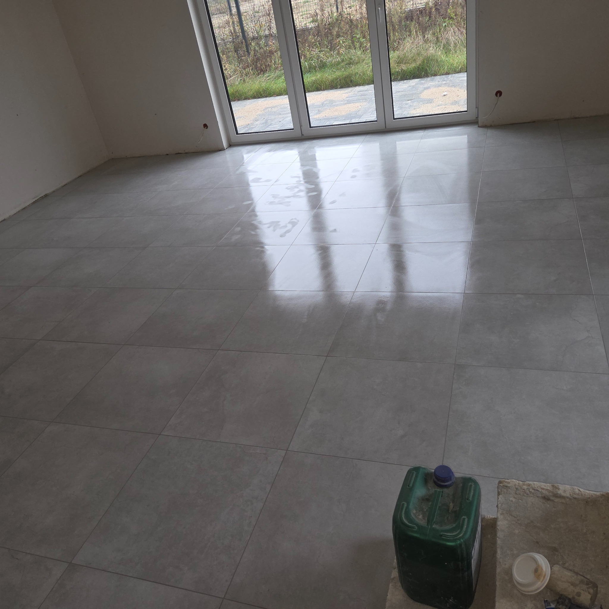 Wykończona podłoga z szarych, błyszczących płytek w nowym domu. Widoczne duże okno balkonowe i pojemnik z płynem do czyszczenia. Minimalistyczny styl.