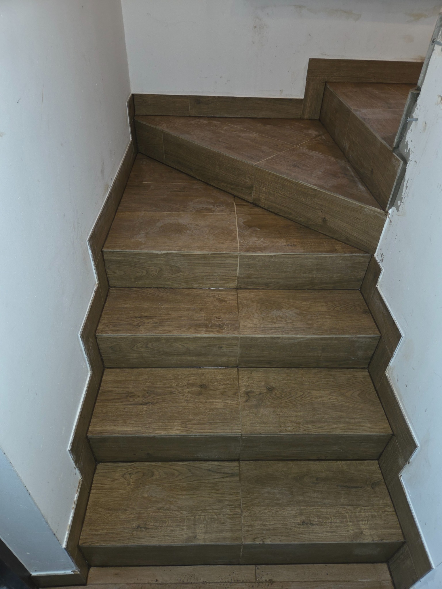 Wykończone schody z płytek imitujących drewno, widok z góry. Białe ściany po bokach. Detal wykończenia krawędzi płytek. Układ schodów w kształcie zygzaka.