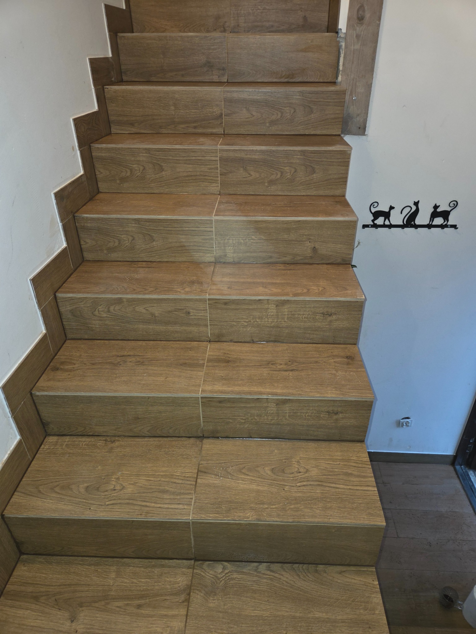 Schody wyłożone brązowymi płytkami imitującymi drewno, z dekoracją ścienną w postaci sylwetek kotów. Widok z dołu, podkreślający wzór płytek i precyzję wykonania.