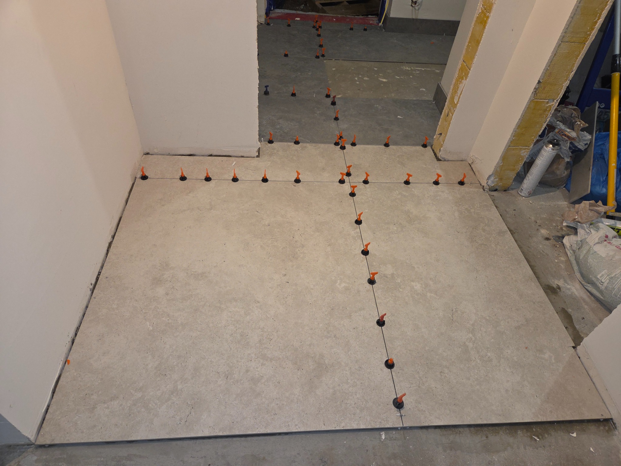 Układanie jasnych płytek podłogowych z użyciem systemu poziomowania, widoczne czarne klipsy i pomarańczowe kliny. Fragment ściany i przygotowane podłoże w tle.