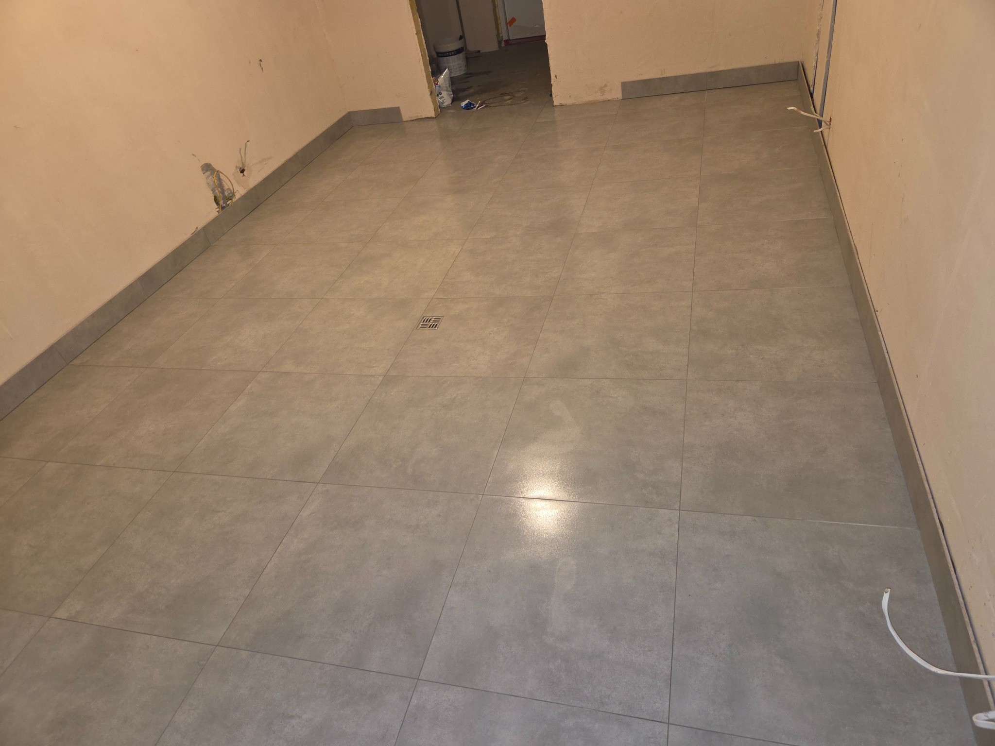 Ułożone szare płytki podłogowe w pomieszczeniu w trakcie remontu. Widoczny odpływ liniowy i wystające kable ze ściany. Płytki ułożone równo, z zachowaniem fug.