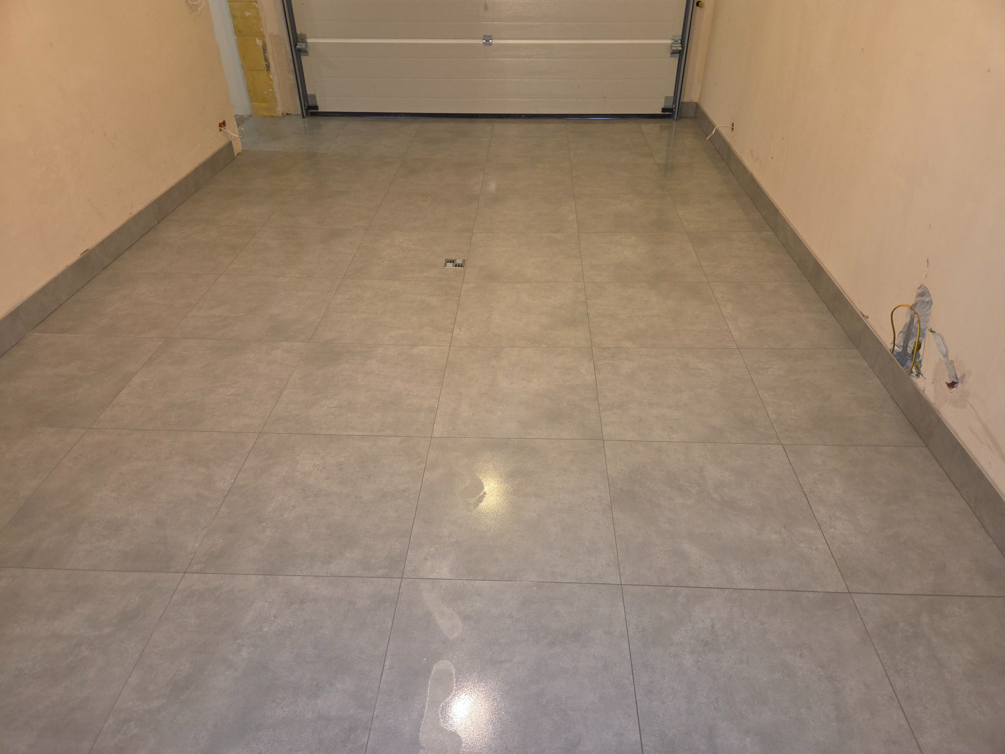 Wykończona podłoga w garażu z szarych, kwadratowych płytek. Widoczny odpływ liniowy. Ściany w kolorze beżowym, surowe wykończenie. Brama garażowa w tle.