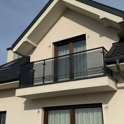 MastMet - Nowoczesny balkon z czarną ramą i szklanym wypełnieniem, widok z dołu na tle jasnego nieba. Minimalistyczny design, eleganckie wykończenie.