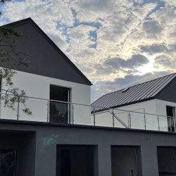 MastMet - Nowoczesny dom z balkonem zabezpieczonym szkłem, widok z dołu na tle pochmurnego nieba. Minimalistyczny design, szklane panele balustrady.
