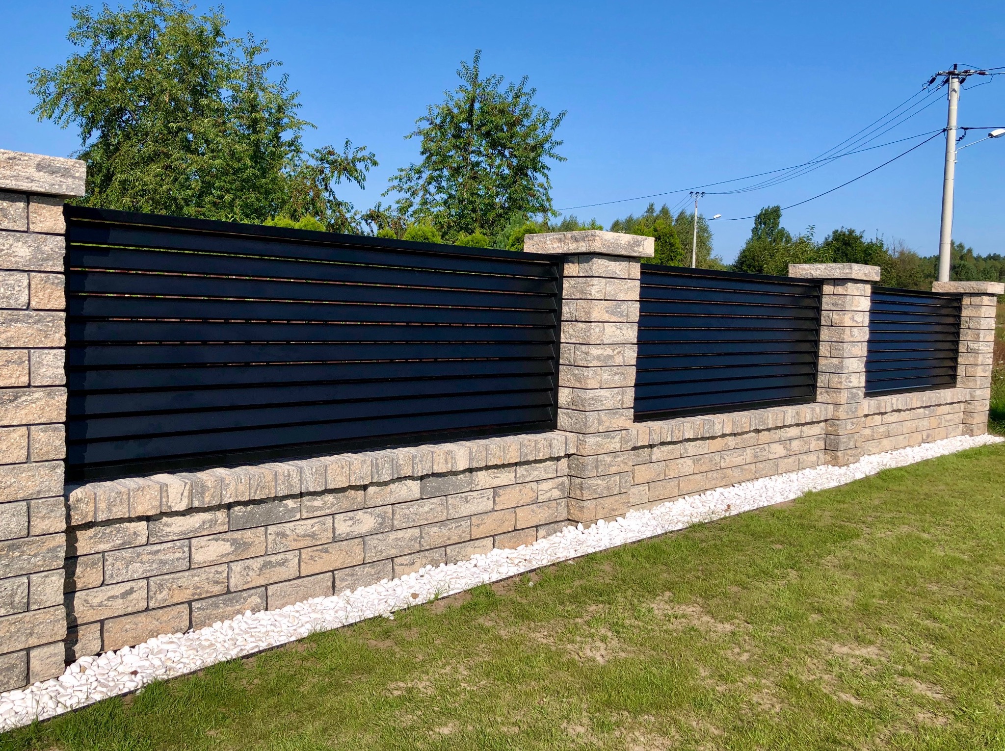 Nowoczesne ogrodzenie z czarnych, poziomych paneli i kamiennych słupków, otoczone białym żwirem i zielonym trawnikiem na tle błękitnego nieba.