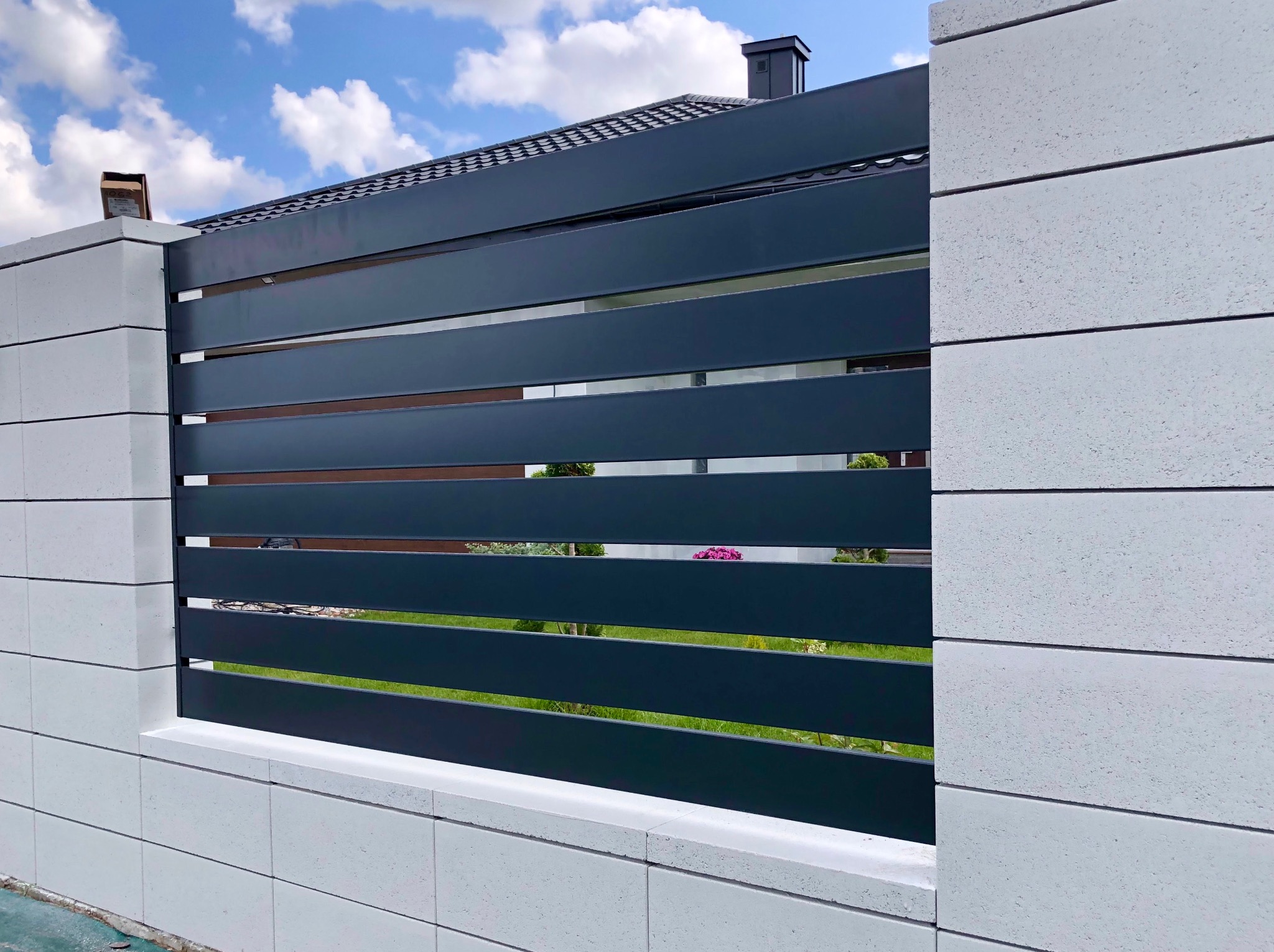 Nowoczesne ogrodzenie z poziomych, grafitowych paneli na białych, betonowych słupkach. Widok na zielony trawnik i dom przez prześwity. Błękitne niebo z chmurami w tle.