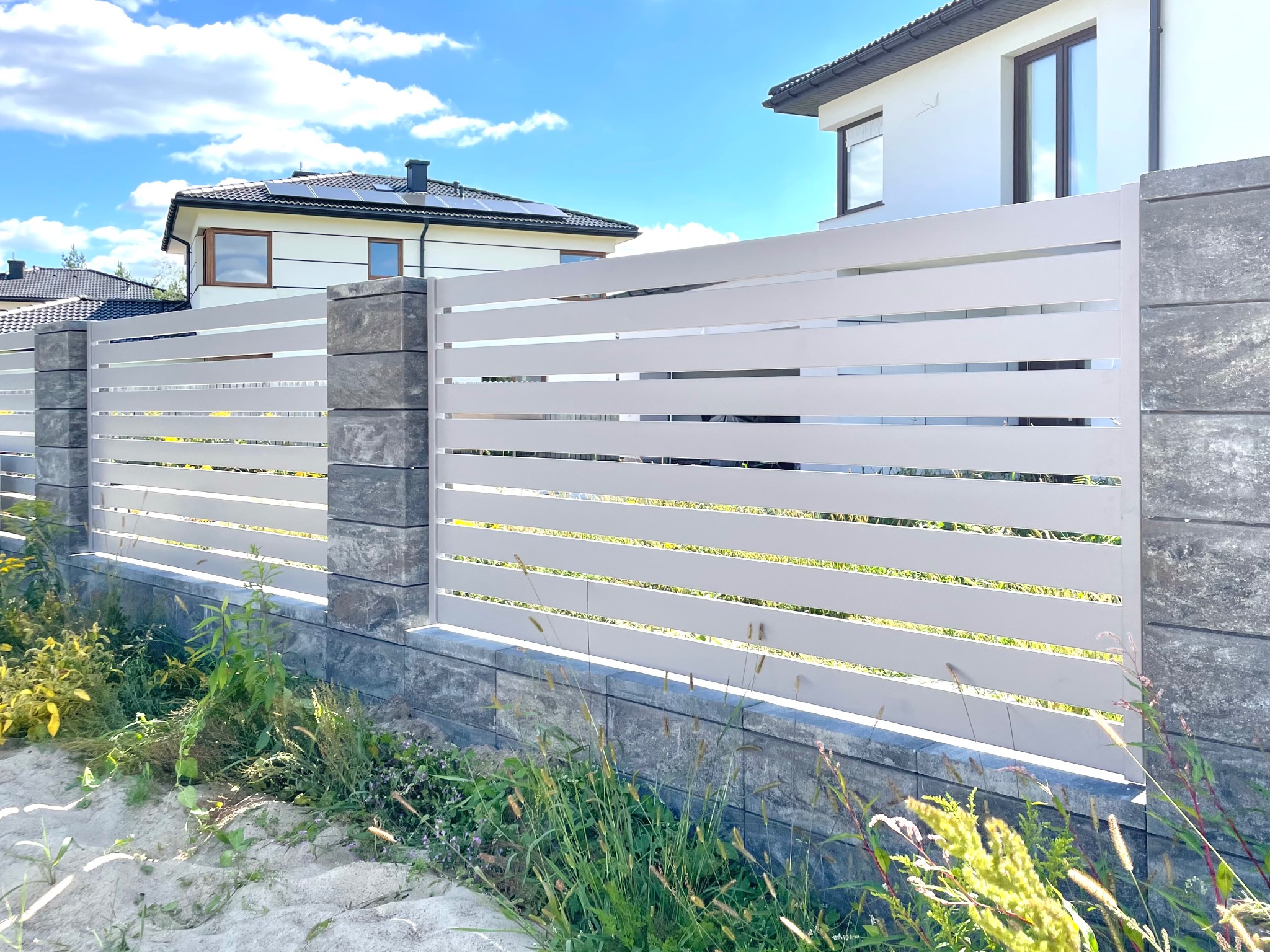 Nowoczesne ogrodzenie posesji z poziomych, białych paneli, wsparte na szarych, kamiennych słupkach. W tle dom z panelami słonecznymi i błękitne niebo.