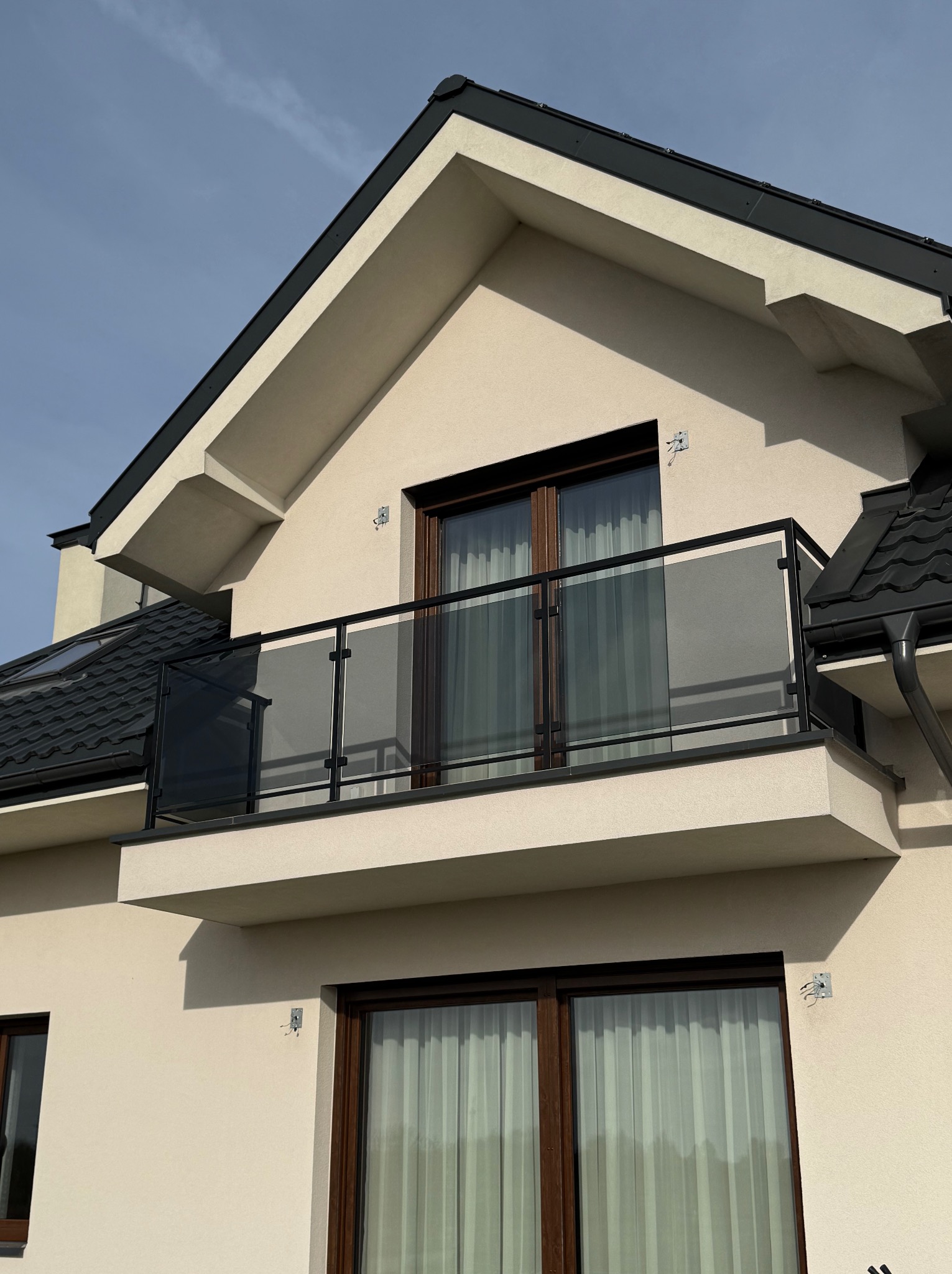 Nowoczesny balkon z czarną ramą i szklanym wypełnieniem, widok z dołu na tle jasnego nieba. Minimalistyczny design, eleganckie wykończenie.
