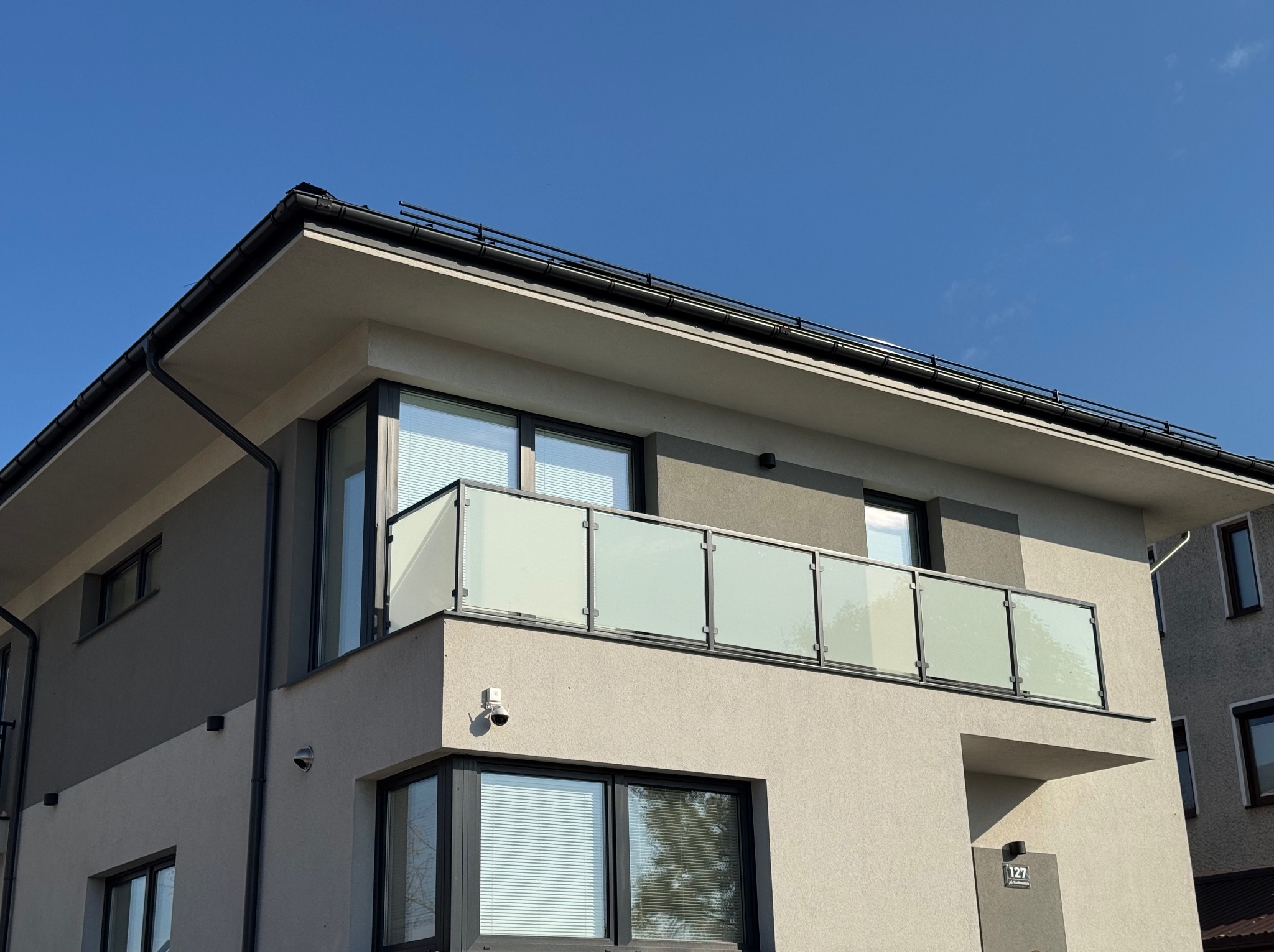 Nowoczesny balkon z szklaną balustradą w budynku o minimalistycznej architekturze, oświetlony jasnym, naturalnym światłem słonecznym. Widoczny numer domu 127.