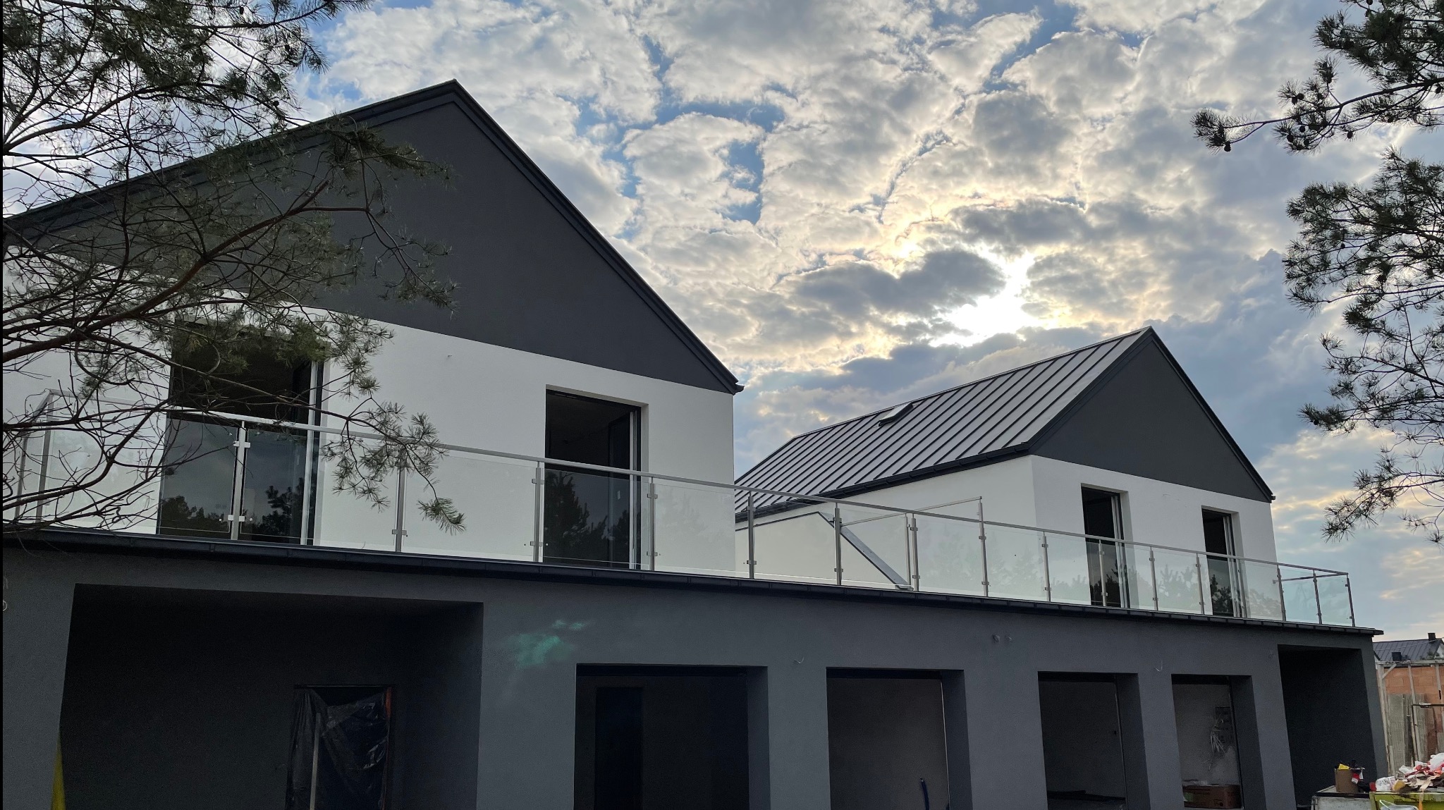 Nowoczesny dom z balkonem zabezpieczonym szkłem, widok z dołu na tle pochmurnego nieba. Minimalistyczny design, szklane panele balustrady.