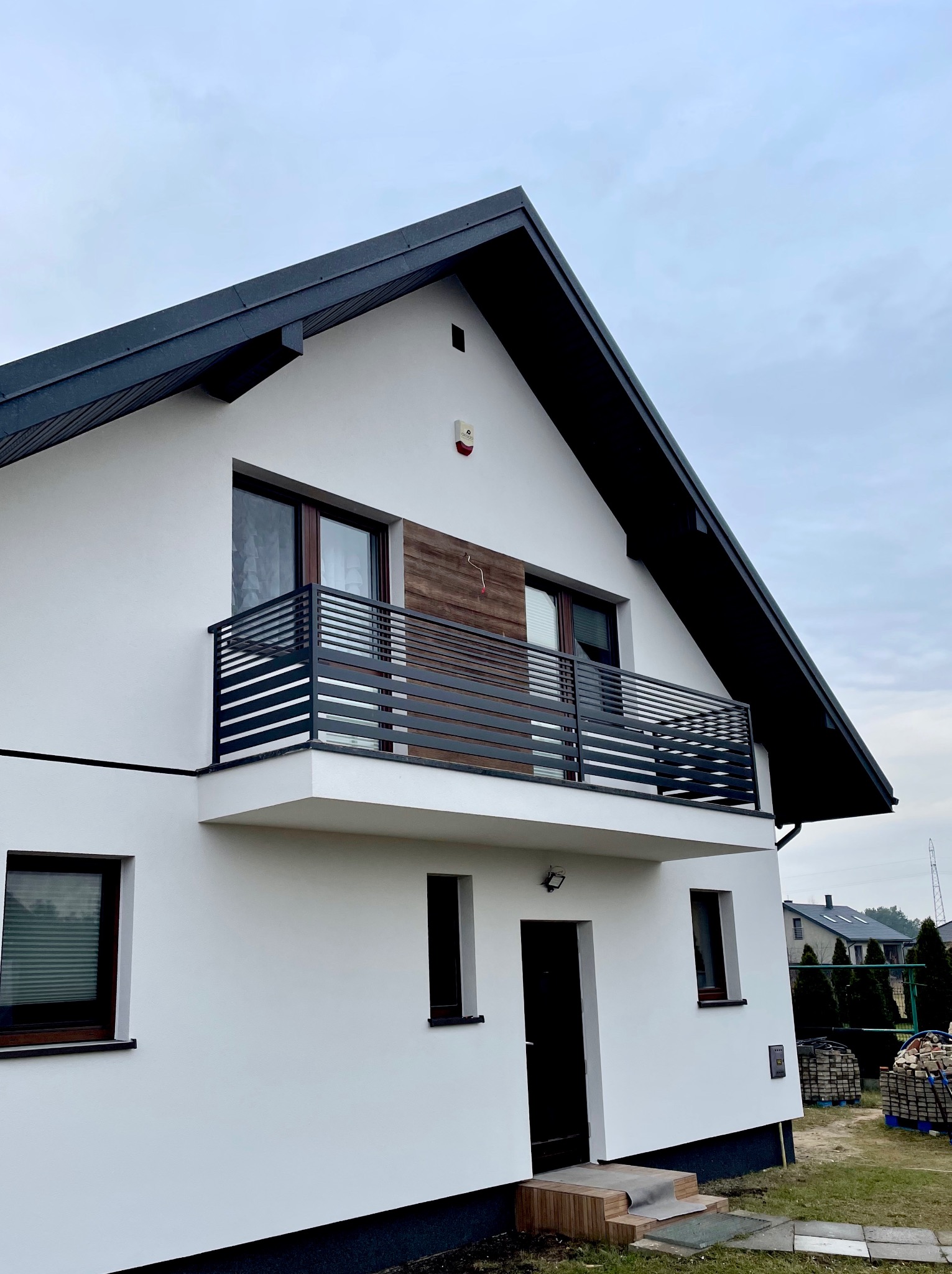 Nowoczesny dom z balkonem i czarną, poziomą balustradą. Biała elewacja, ciemne okna i dach. Widok z dołu, pochmurne niebo w tle. Minimalistyczny design.