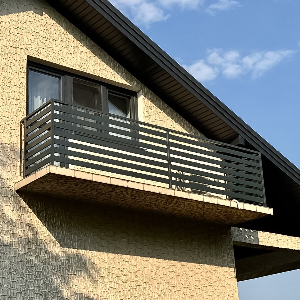 Nowoczesny balkon z czarną, poziomą balustradą metalową, widoczny z dołu pod kątem. Jasna elewacja budynku i fragment błękitnego nieba w tle. Minimalistyczny design.