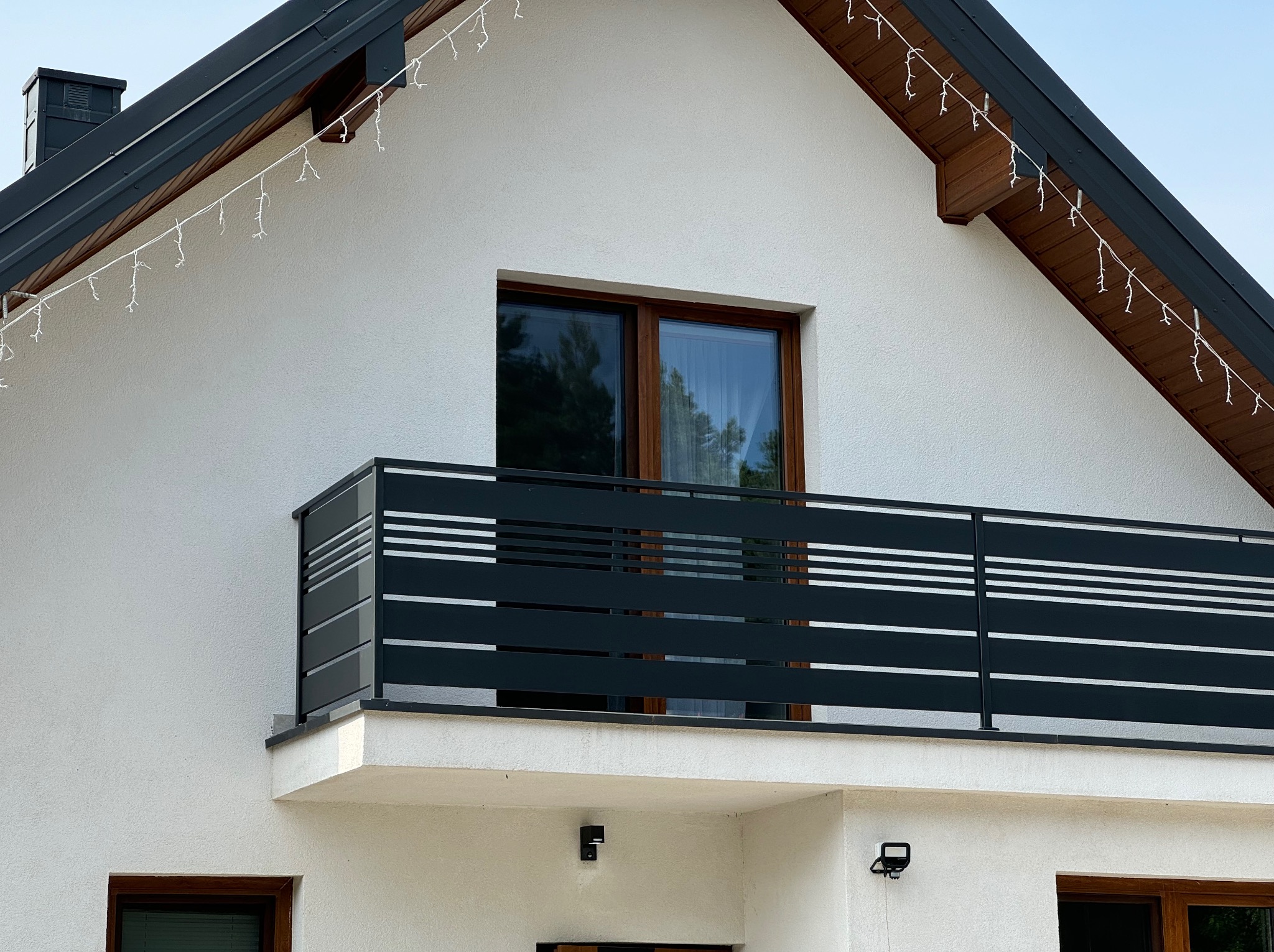 Czarna, pozioma balustrada balkonowa z jasnymi paskami na tle białej elewacji domu z oknem i świątecznymi lampkami.