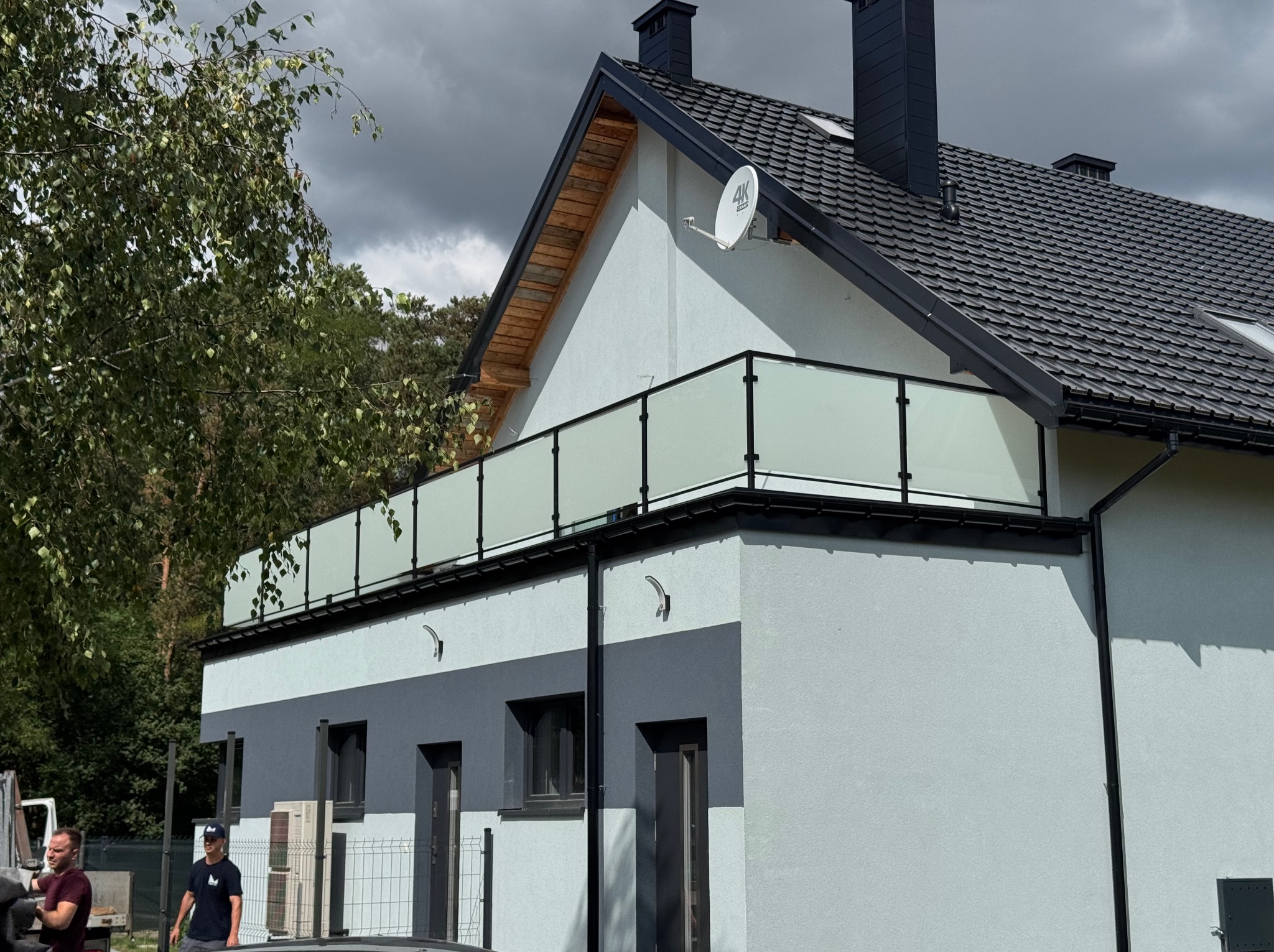 Nowoczesny dom z balkonem zabezpieczonym szklaną balustradą. Widoczne czarne ramy i przezroczyste szkło. Na dachu antena satelitarna.