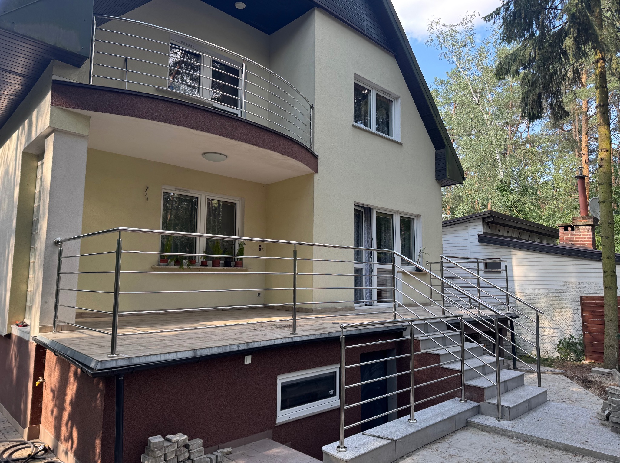 Dwupoziomowy dom z balkonem i tarasem. Stalowe, poziome barierki zabezpieczają przestrzeń. Widoczne schody wejściowe i fragment ogrodu z drzewami.