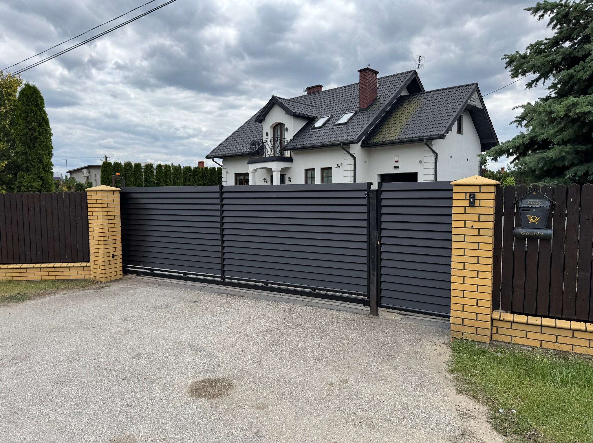 Nowoczesna, pozioma brama metalowa w kolorze antracytowym, z ceglanymi słupkami, na tle białego domu z ciemnym dachem i pochmurnego nieba. Skrzynka na listy.
