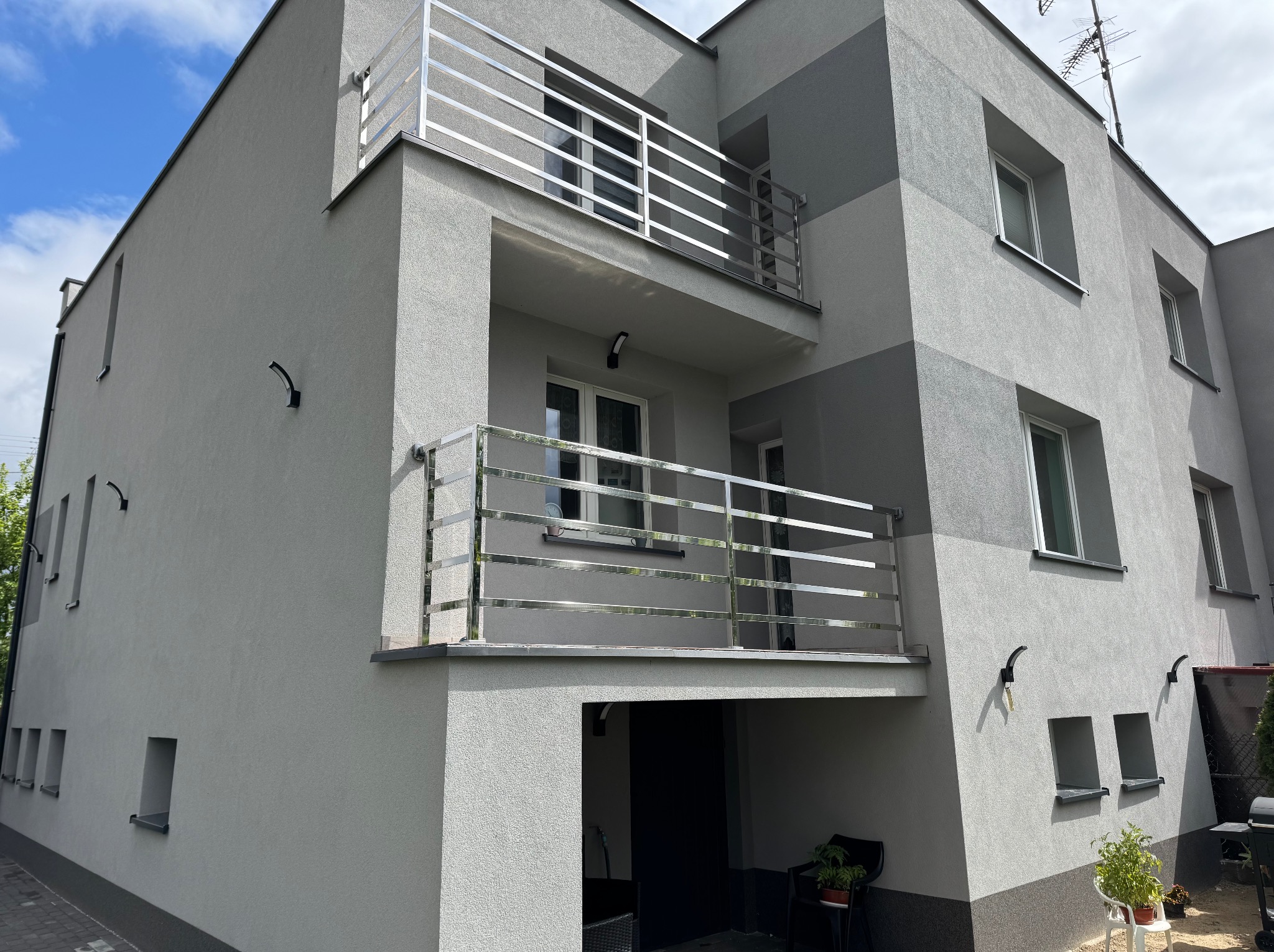 Nowoczesny dom z balkonami zabezpieczonymi prostymi, poziomymi, metalowymi barierkami. Szara elewacja, minimalistyczny design, rośliny w donicach.