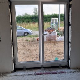 SAM-DREW Kamil Samuś - Białe, dwuskrzydłowe okno PCV z roletą zewnętrzną w surowym otworze ściany. Widok na zewnątrz: samochód, cegły i zieleń. Wnętrze w stanie deweloperskim.