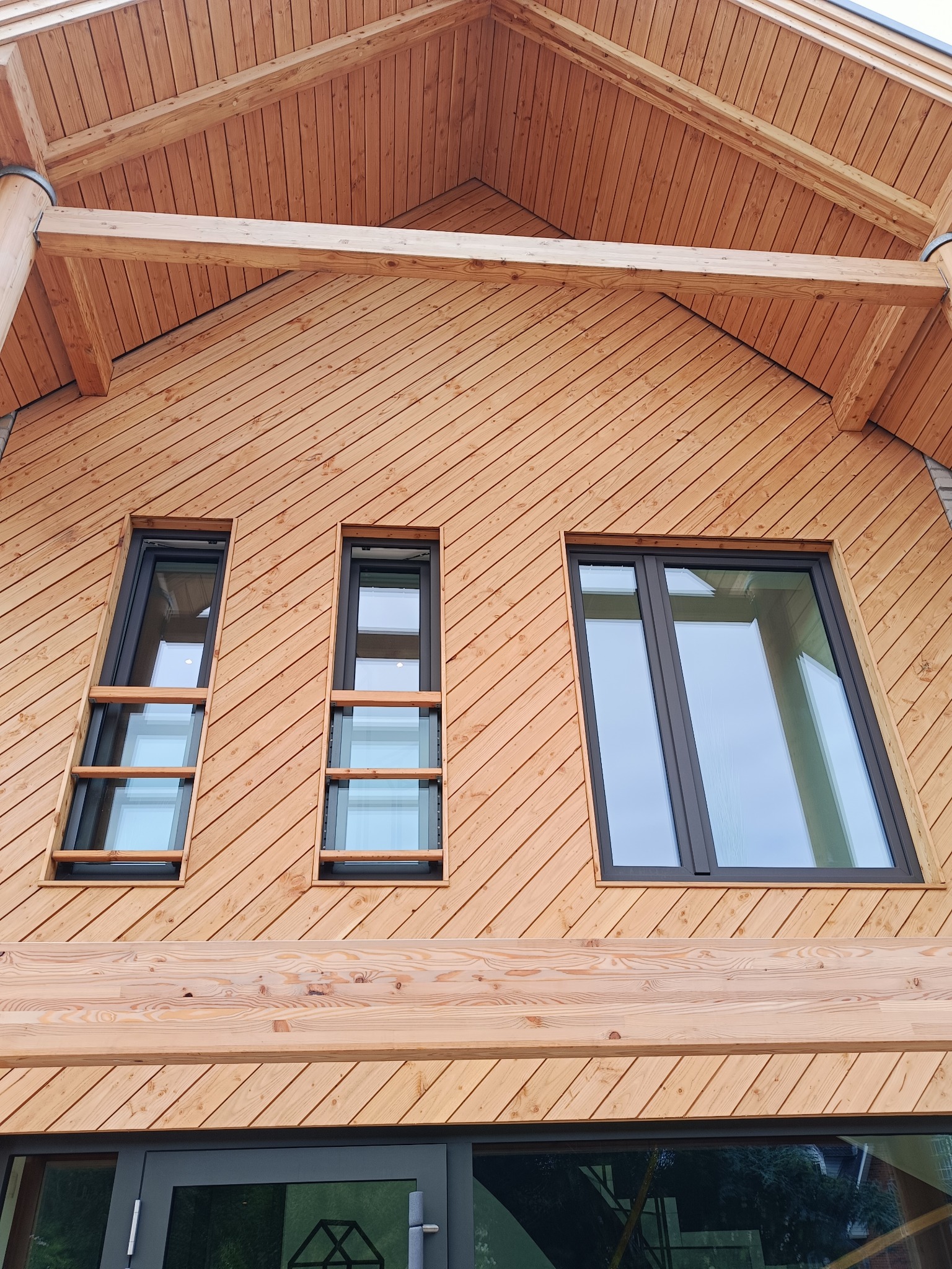 Elewacja domu z drewnianą boazerią i trzema oknami PCV w czarnych ramach. Ujęcie z dołu, widoczne elementy konstrukcyjne dachu i drzwi wejściowe.