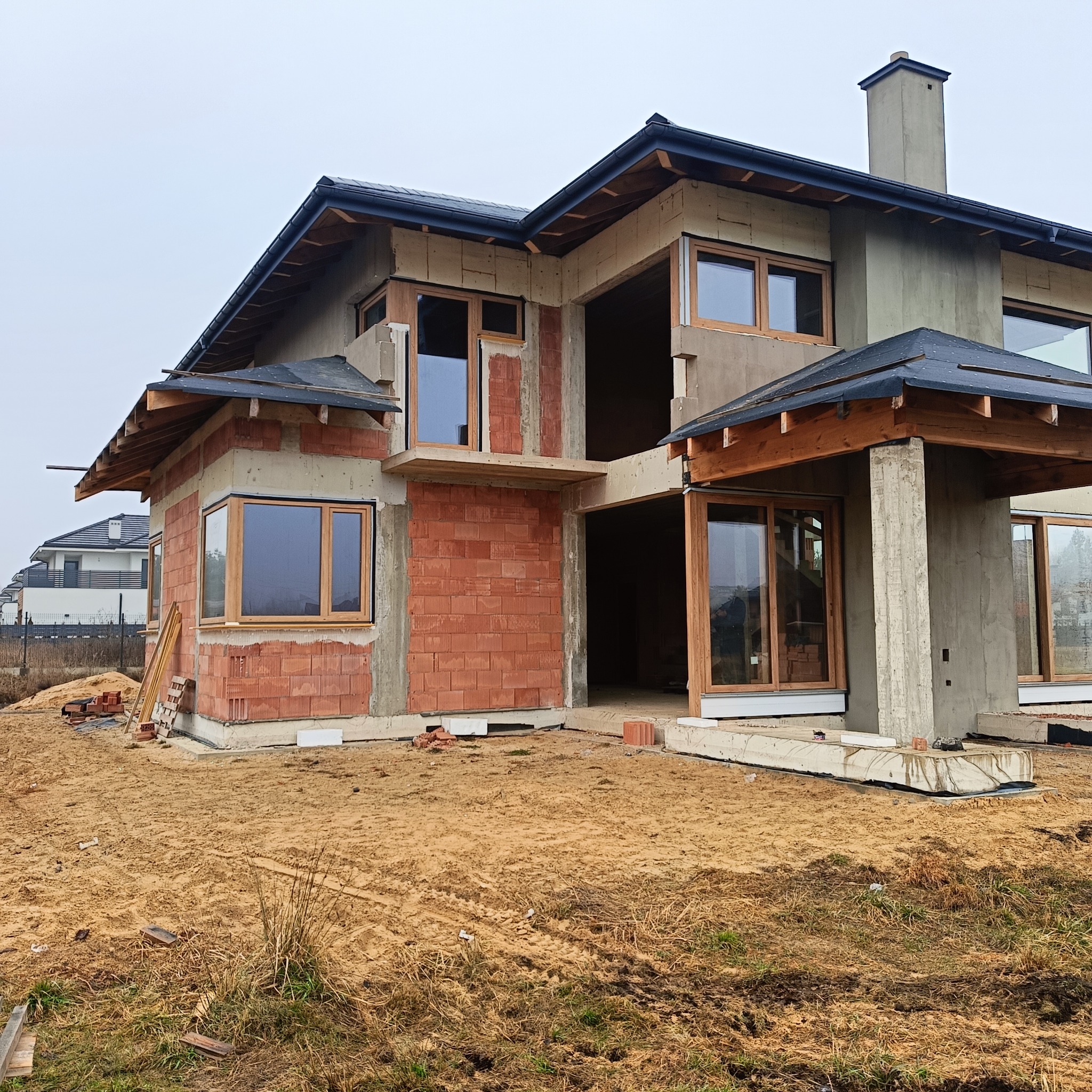 Surowy stan domu jednorodzinnego z zamontowanymi oknami z drewnianymi ramami. Widoczne cegły, beton i elementy konstrukcji dachu. Teren wokół w stanie budowy.