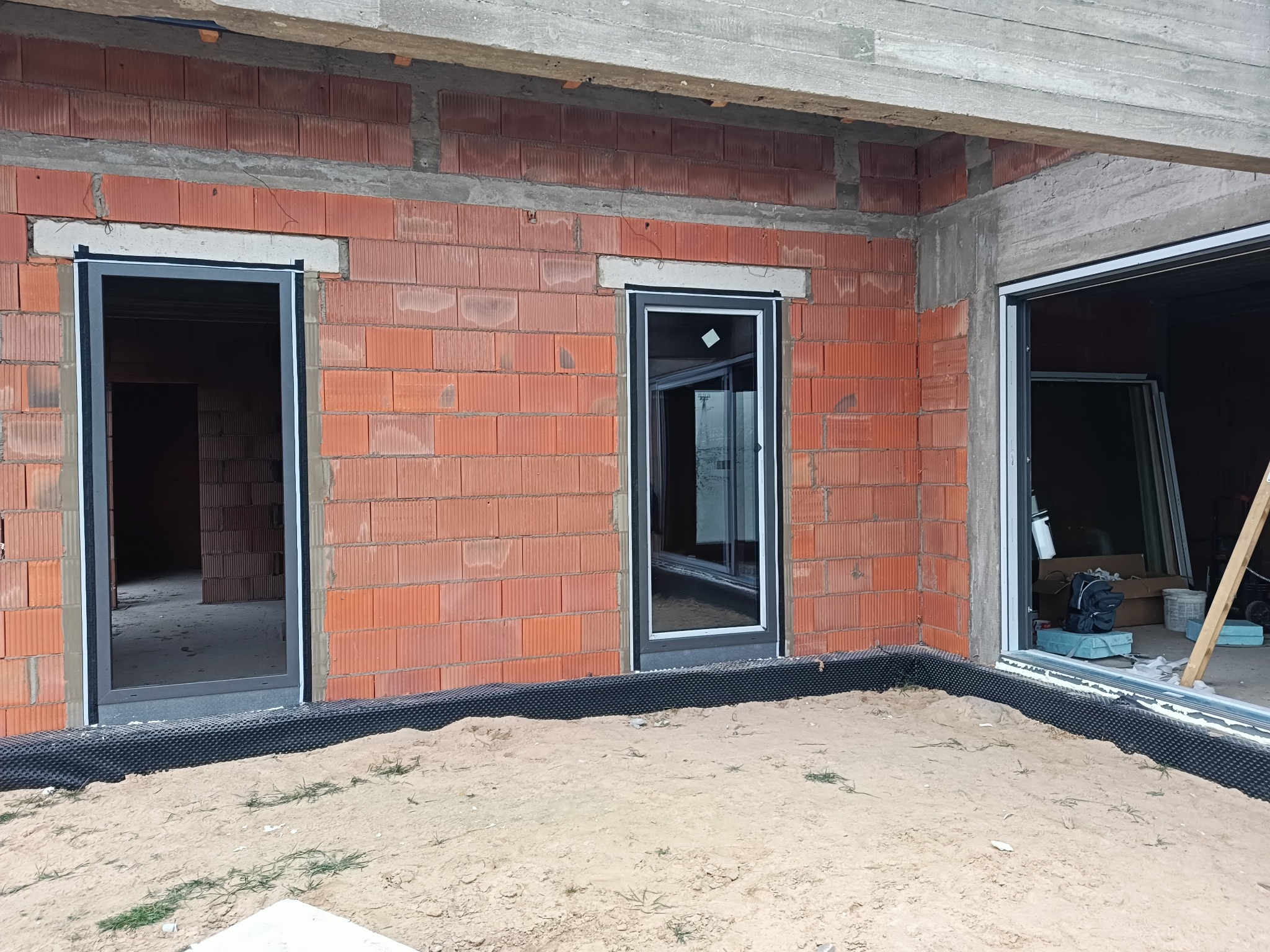 Surowy stan: wstawione ramy okienne i drzwiowe w murze z czerwonej cegły dziurawki, widoczne detale montażu, piasek na podłożu.