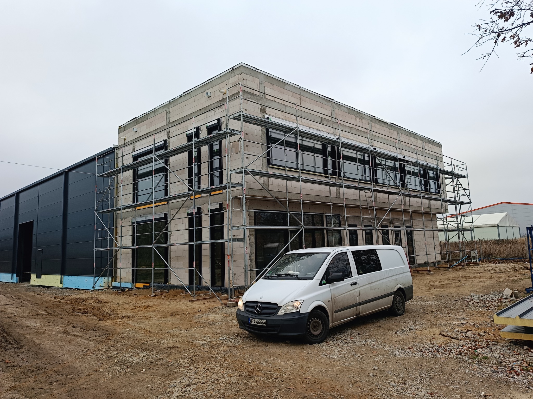 Budynek w trakcie montażu okien, otoczony rusztowaniami. Na pierwszym planie biały samochód dostawczy. Widoczne elementy izolacji. Pochmurne niebo w tle.