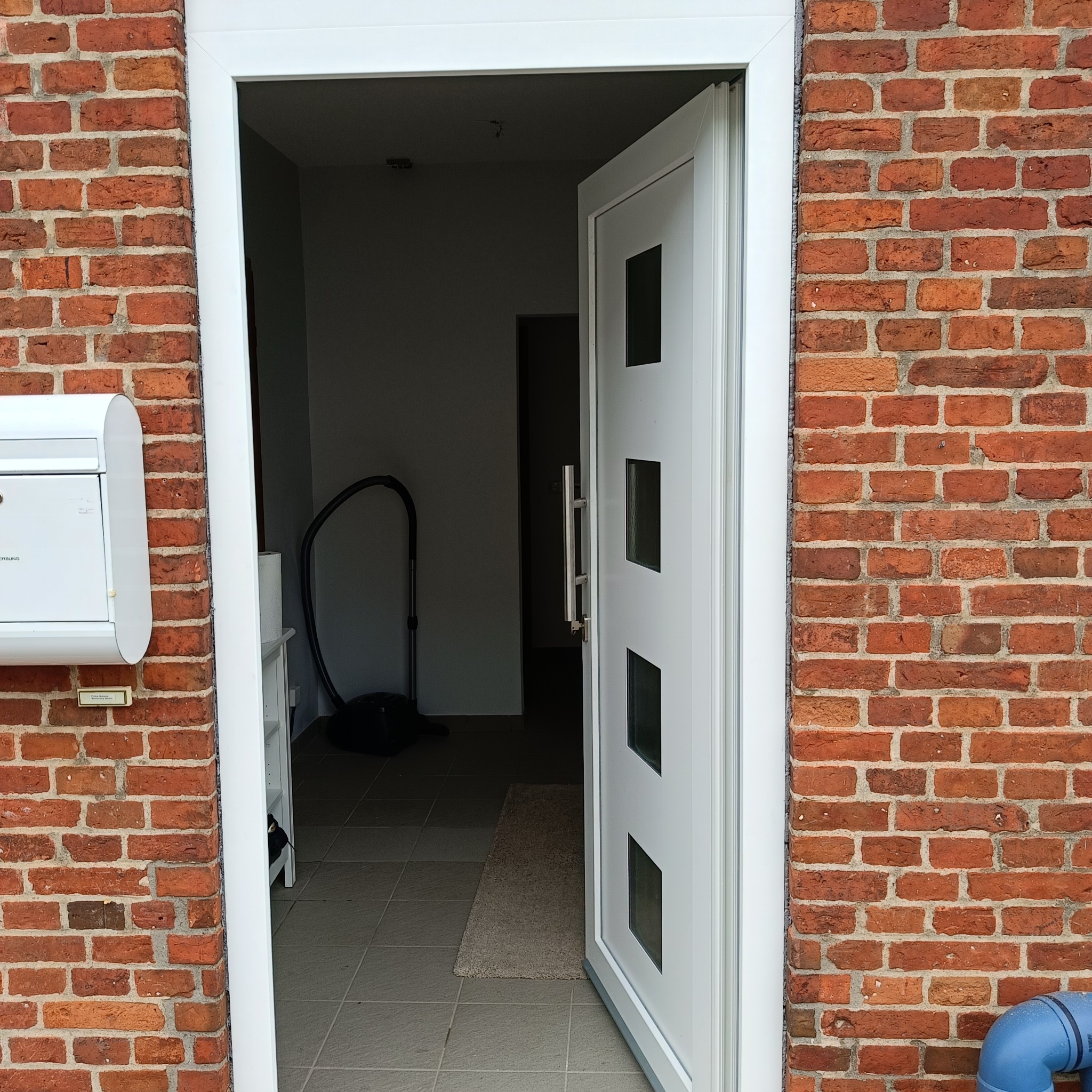Otwarta, biała, nowoczesna drzwi wejściowe z czterema szybkami, zamontowana w murze z czerwonej cegły. Widoczna skrzynka pocztowa i fragment wnętrza.