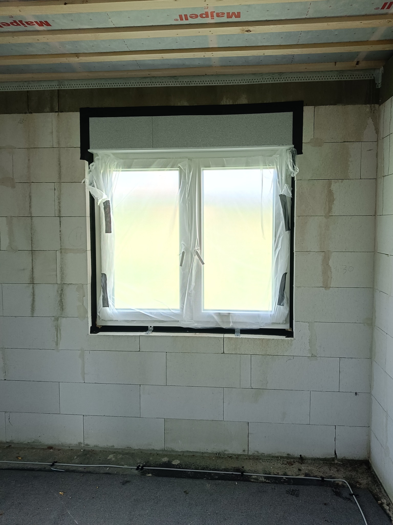 Nowe okno PCV, zabezpieczone folią, osadzone w surowej ścianie z betonu komórkowego. Widoczne czarne uszczelnienie wokół ramy i drewniana konstrukcja stropu.