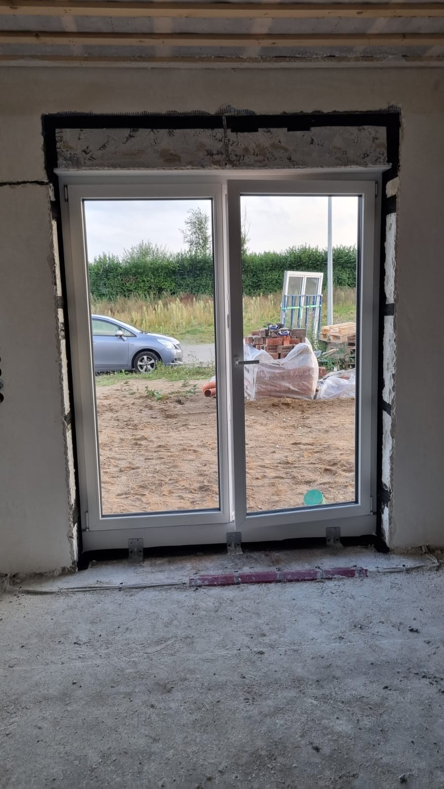 Białe, dwuskrzydłowe okno PCV z roletą zewnętrzną w surowym otworze ściany. Widok na zewnątrz: samochód, cegły i zieleń. Wnętrze w stanie deweloperskim.
