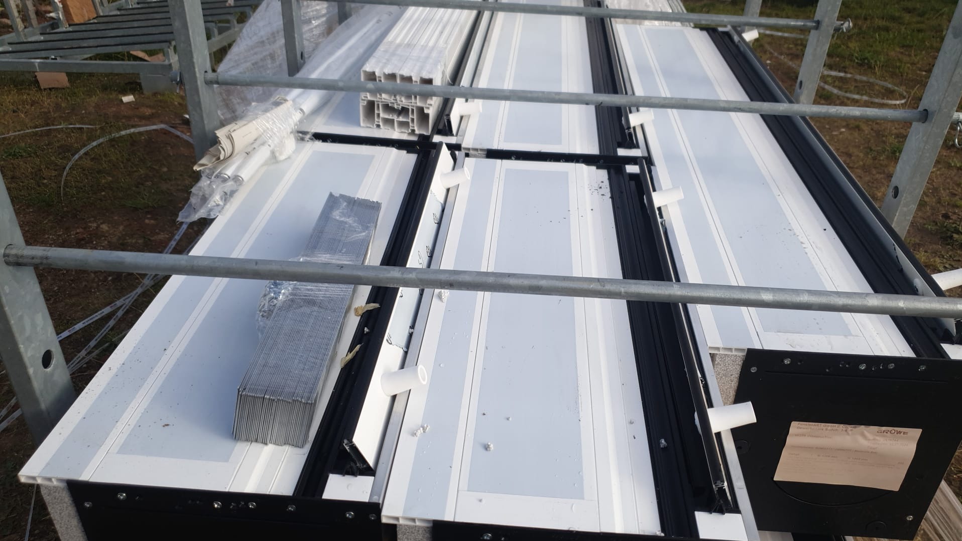Przygotowane do montażu białe ramy okienne PCV z czarnymi uszczelkami, ułożone na metalowym rusztowaniu. Widoczne akcesoria i dokumentacja.
