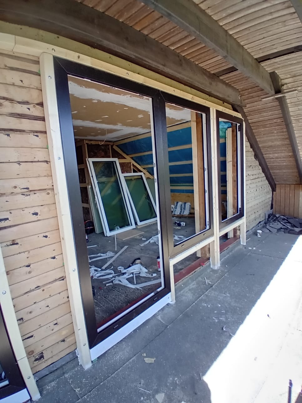 Częściowy montaż okien PCV z czarnymi ramami w drewnianej konstrukcji budynku. W tle widoczne okna czekające na instalację i elementy wykończeniowe.