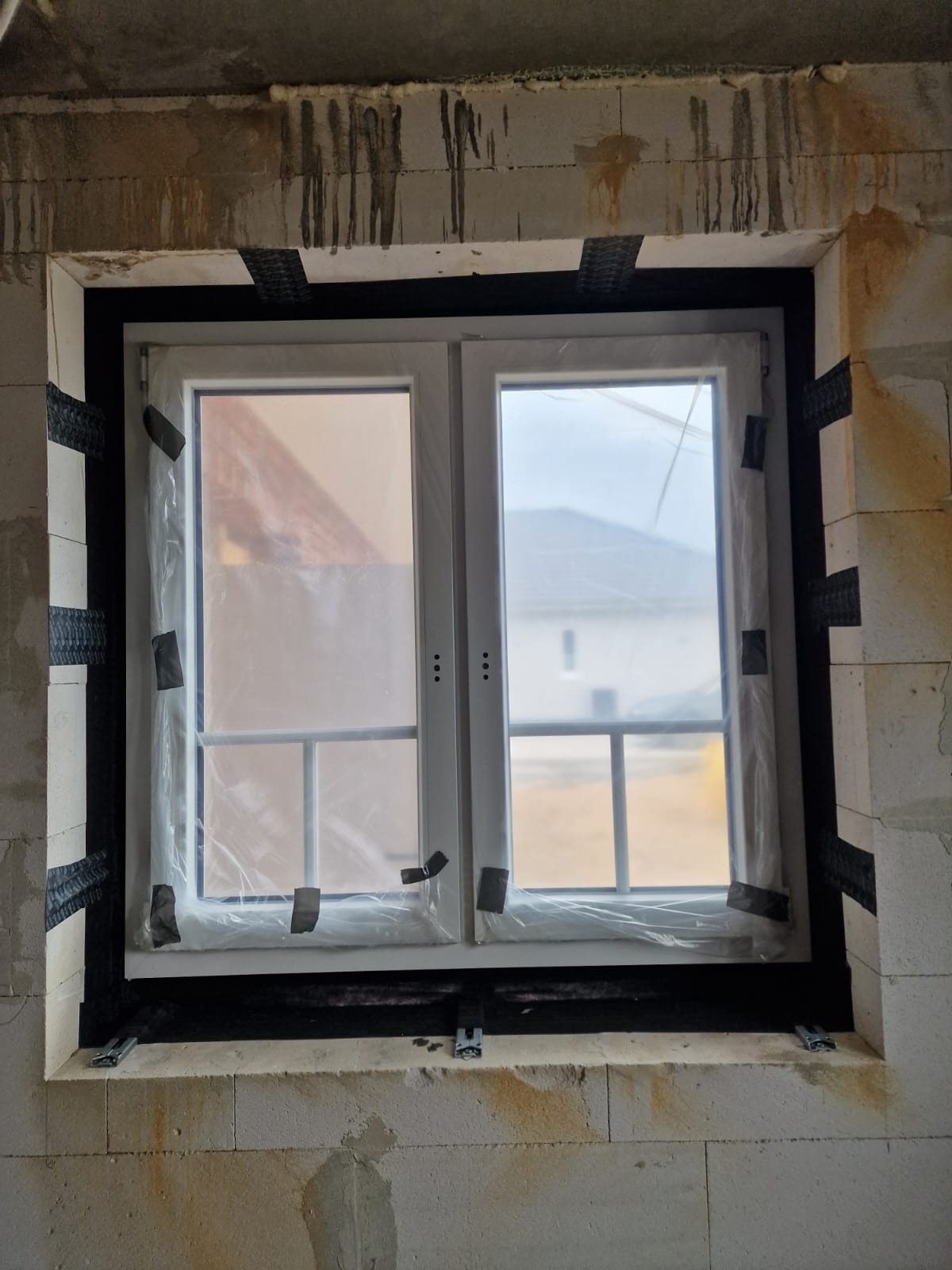 Białe okno PCV z folią ochronną, zamontowane w surowej ścianie z betonu komórkowego. Widoczne taśmy uszczelniające i elementy montażowe.
