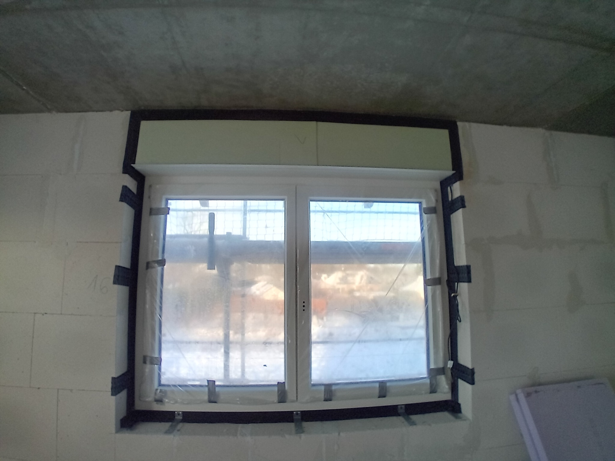 Montaż okna PCV w surowym stanie, zabezpieczone folią i taśmami montażowymi. Widoczne bloczki betonowe i surowy betonowy strop.