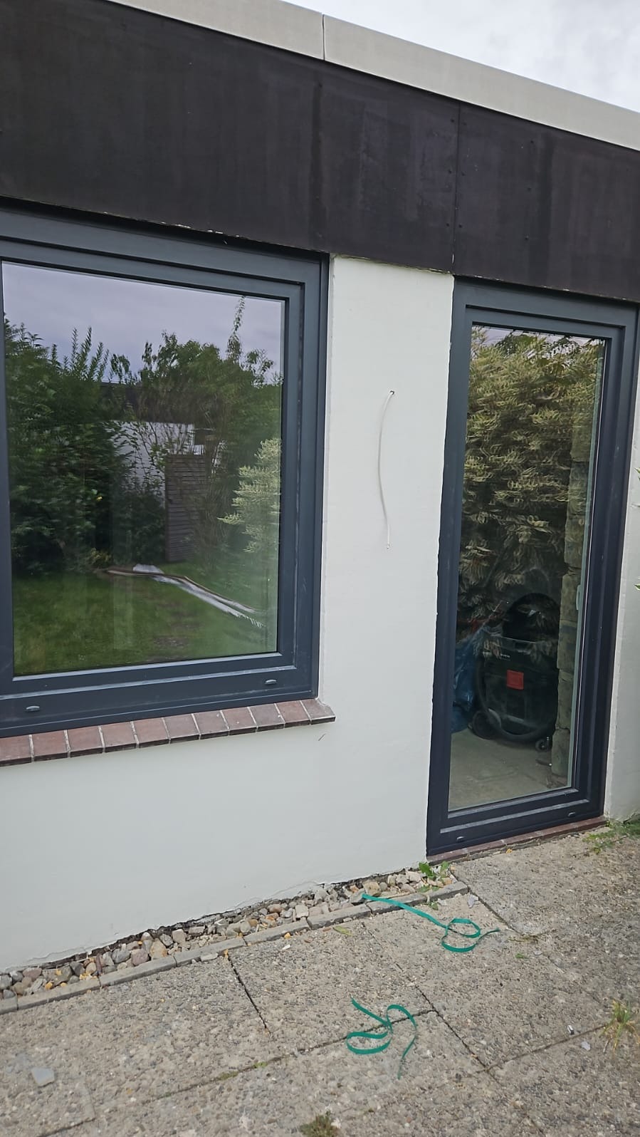 Szare okno PCV i drzwi balkonowe w jasnej elewacji budynku z ciemnym pasem u góry. Widoczne odbicie zieleni w szybach, na zewnątrz kostka brukowa i zwinięty zielony sznurek.