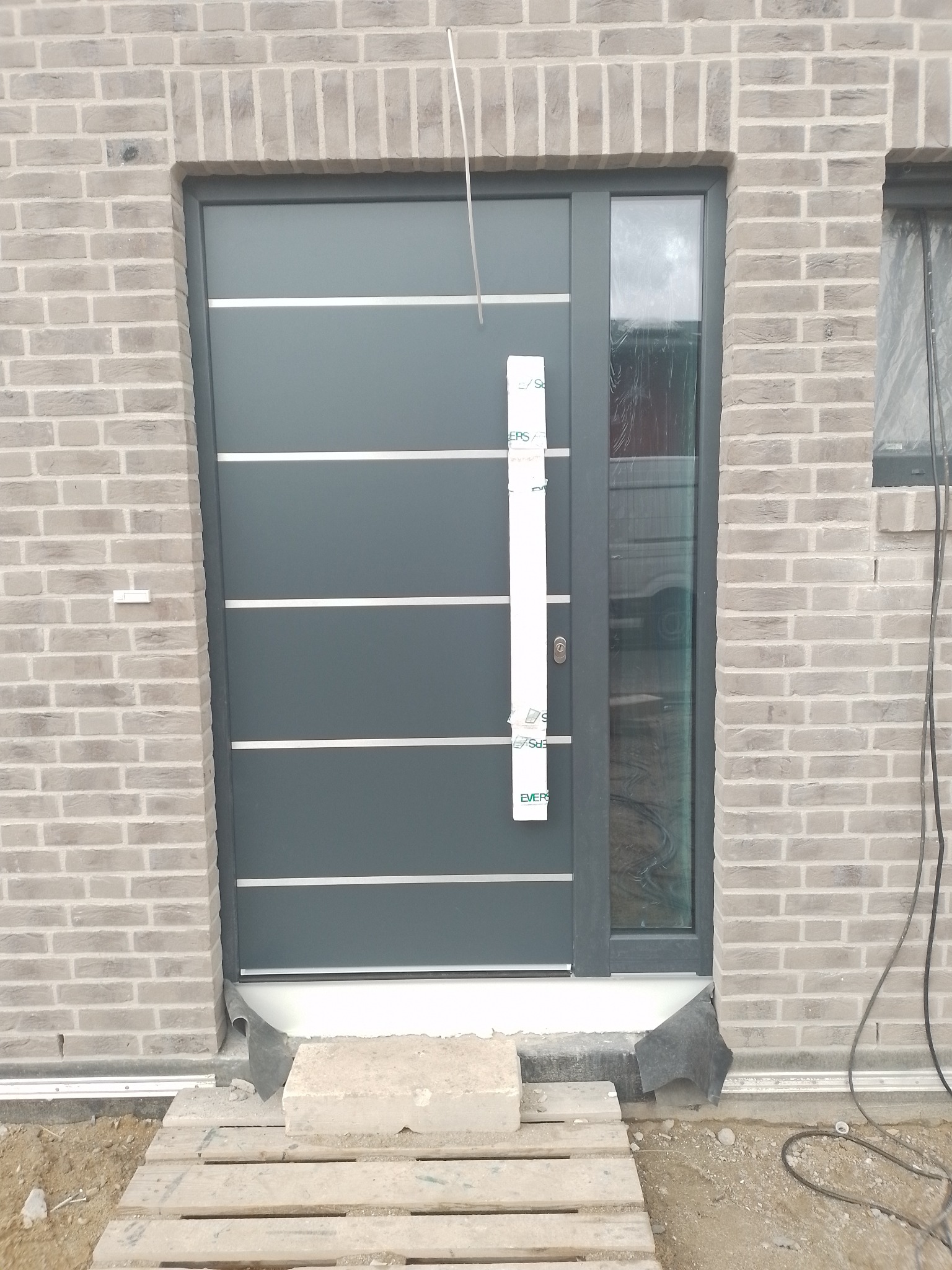 Nowoczesne, grafitowe drzwi wejściowe z poziomymi, srebrnymi akcentami, zamontowane w murze z jasnej cegły. Widoczny fragment okna i tymczasowe zabezpieczenia.